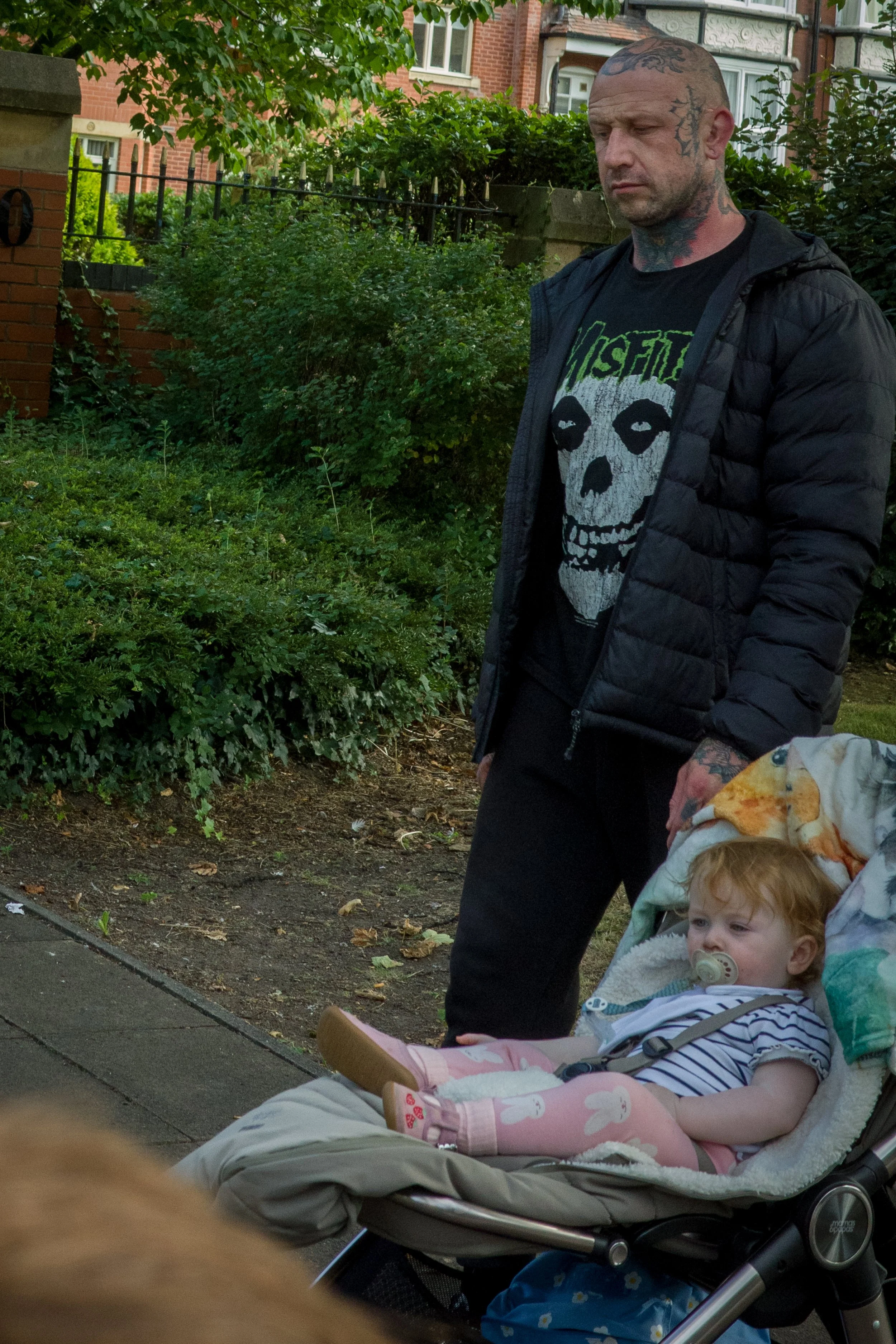 A man with tattoos on his head and neck stands next to a young girl in a stroller outdoors in a city neighborhood with greenery and brick buildings in the background.