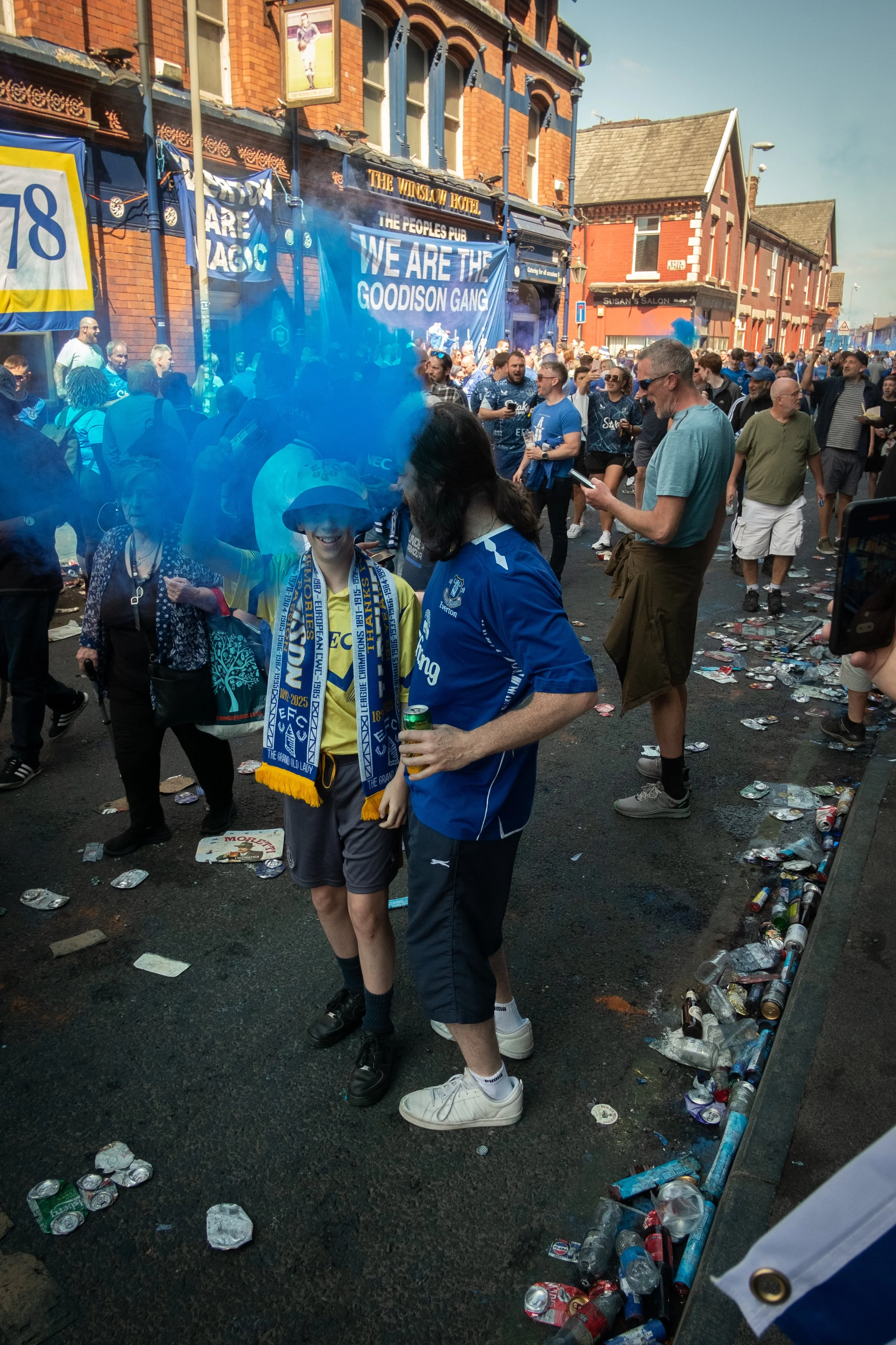 Crowd of football fans celebrating outside a pub with blue smoke, banners, and scattered trash on the street after a match.
