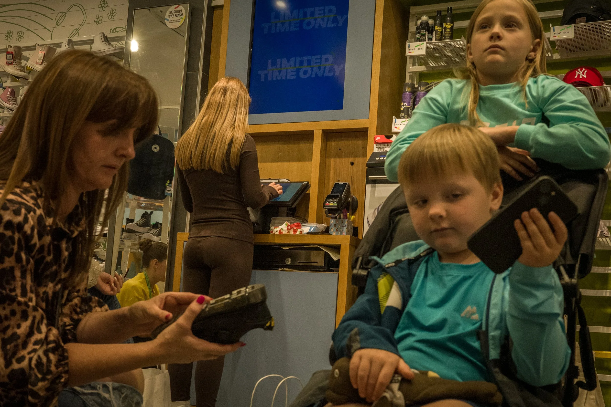 A woman with long brown hair wearing a leopard print top is looking at her phone in a shoe store. Behind her, a man with long blonde hair and a woman with long blonde hair are at the checkout counter, which has a screen and a cash register. To the ri
