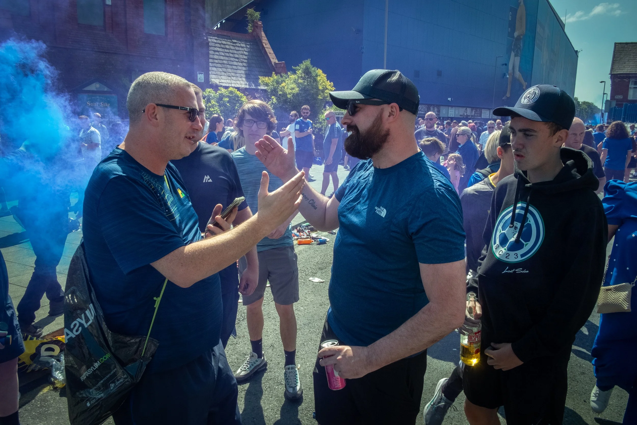 Group of people outdoors during a festival or event, with two men in focus engaging in conversation, one holding a smartphone and the other gesturing with his hand, surrounded by a crowd in casual clothing and colorful smoke.
