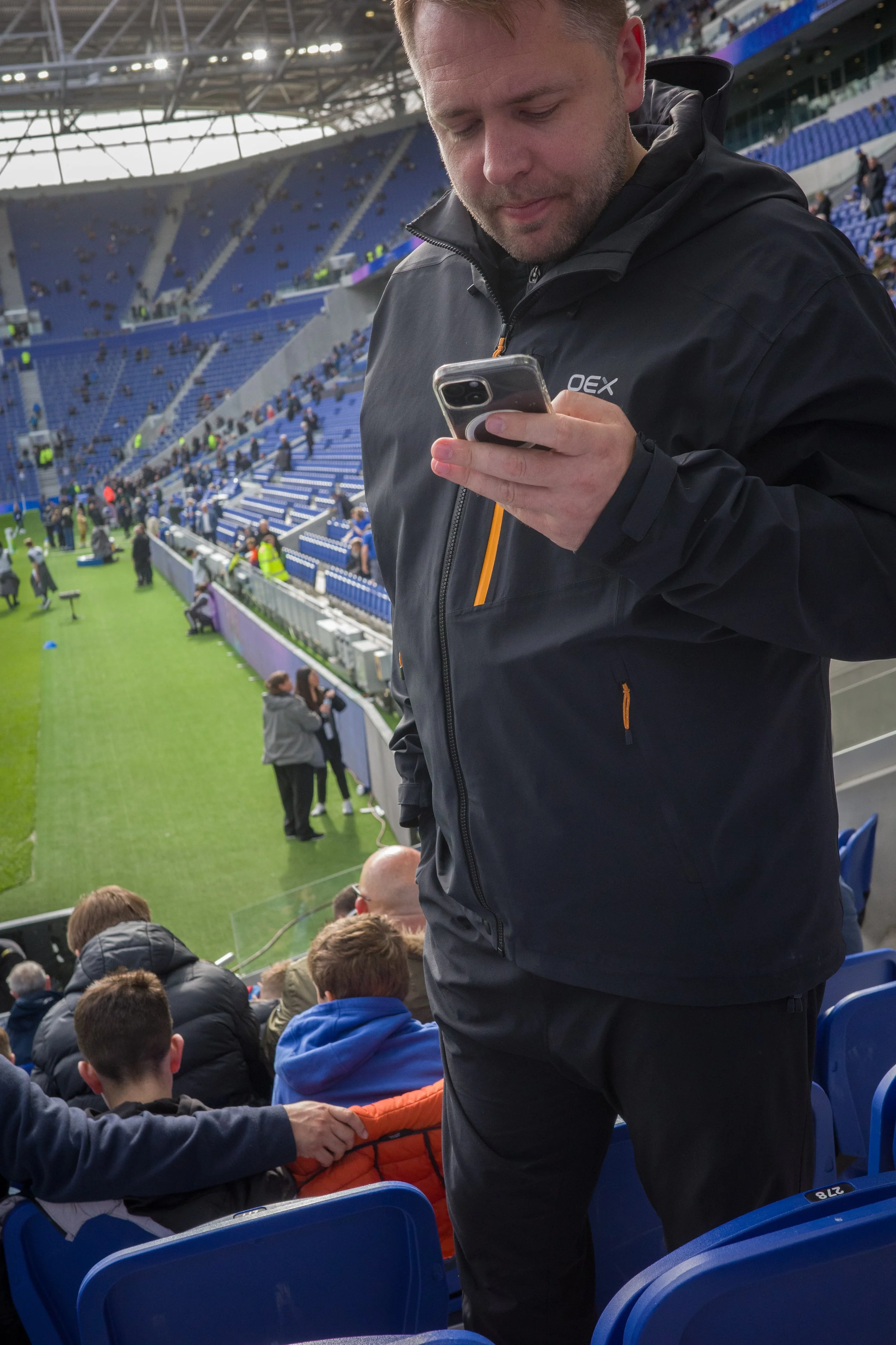 A man wearing a black jacket using a smartphone in a stadium with blue seats. Several people are seated and walking around in the background.