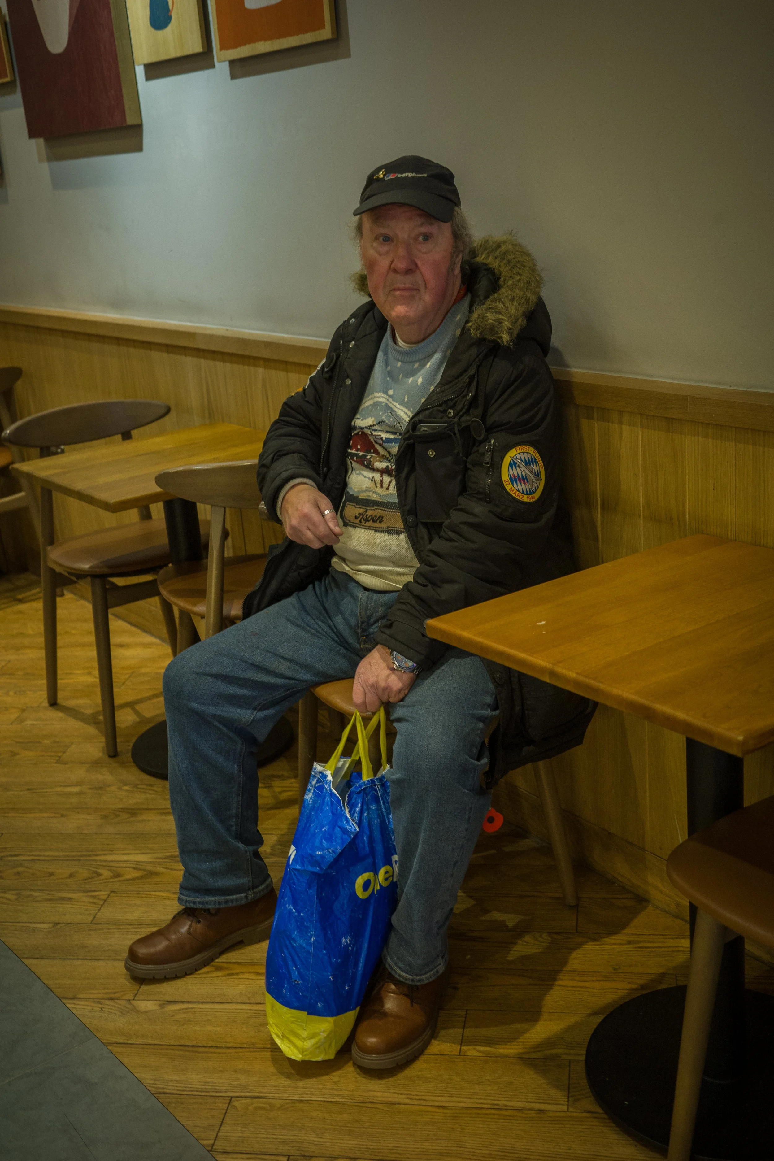 An elderly man sitting on a wooden chair in a restaurant, holding a blue shopping bag. He is wearing a black jacket with a patch on the sleeve, a cap, and jeans.