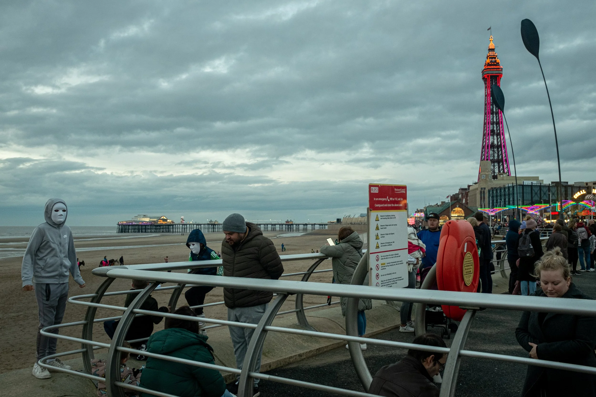 People walking and sitting on benches at a busy beachside pier with an overcast sky, a brightly lit tower, and colorful attractions in the background.