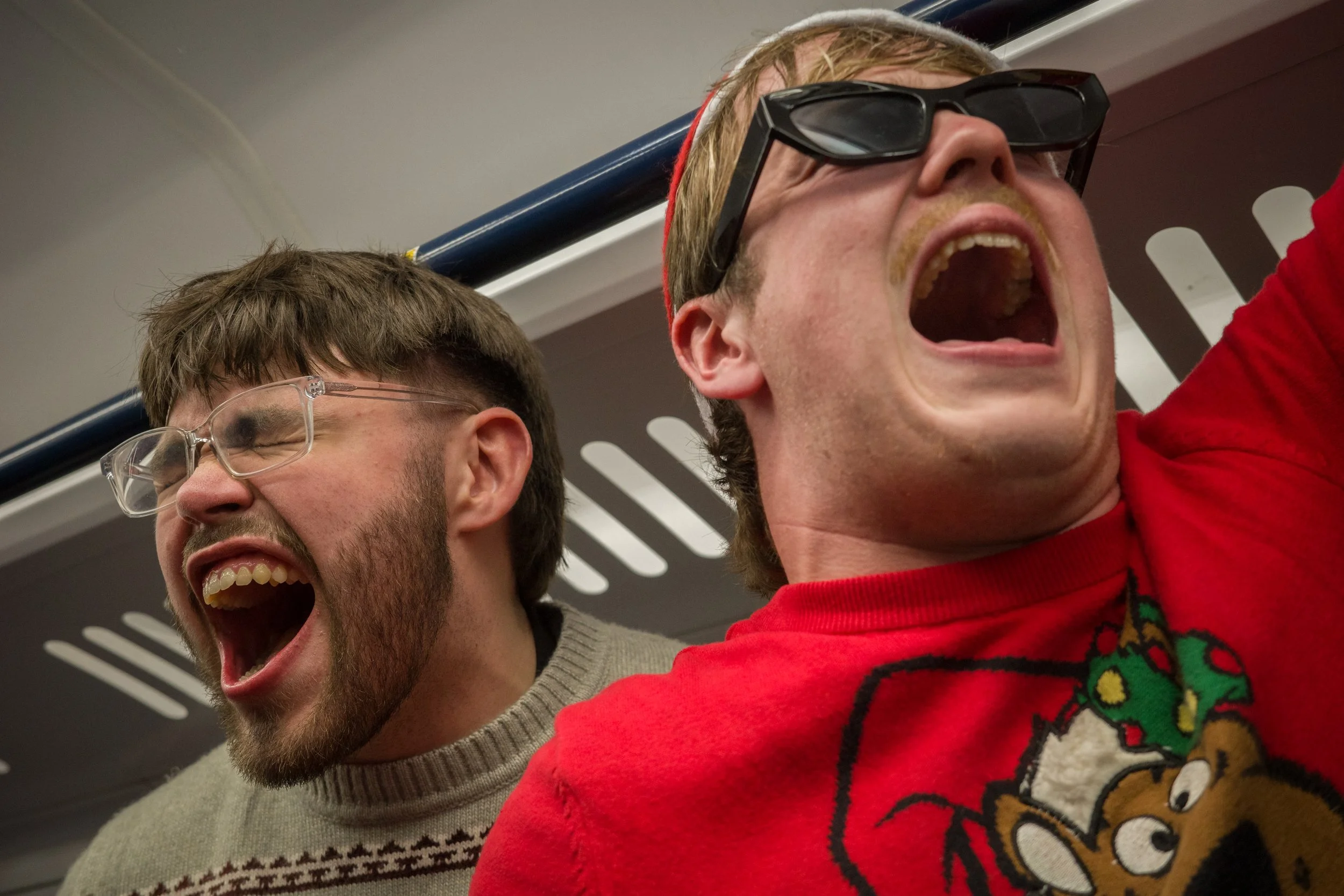 Two men are on a train, both with mouths open as if shouting or singing. The man on the right wears sunglasses and a Christmas sweater with a reindeer, and the man on the left wears glasses and a patterned sweater. They appear excited or animated.
