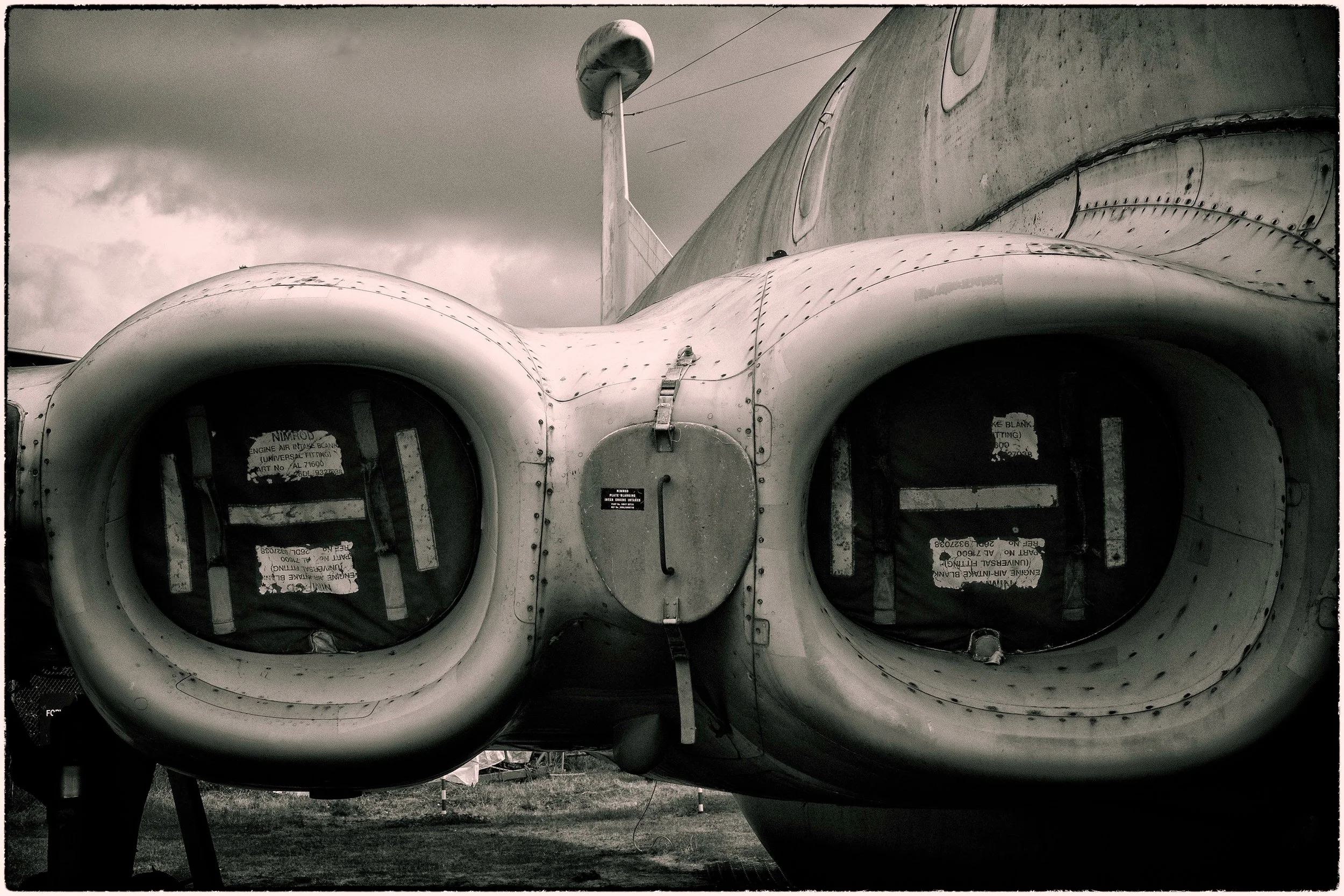 Black and white photograph of a vintage aircraft cockpit viewed from outside. The image shows the front view with large circular windows, rivets along the metal surface, and a background of cloudy sky.