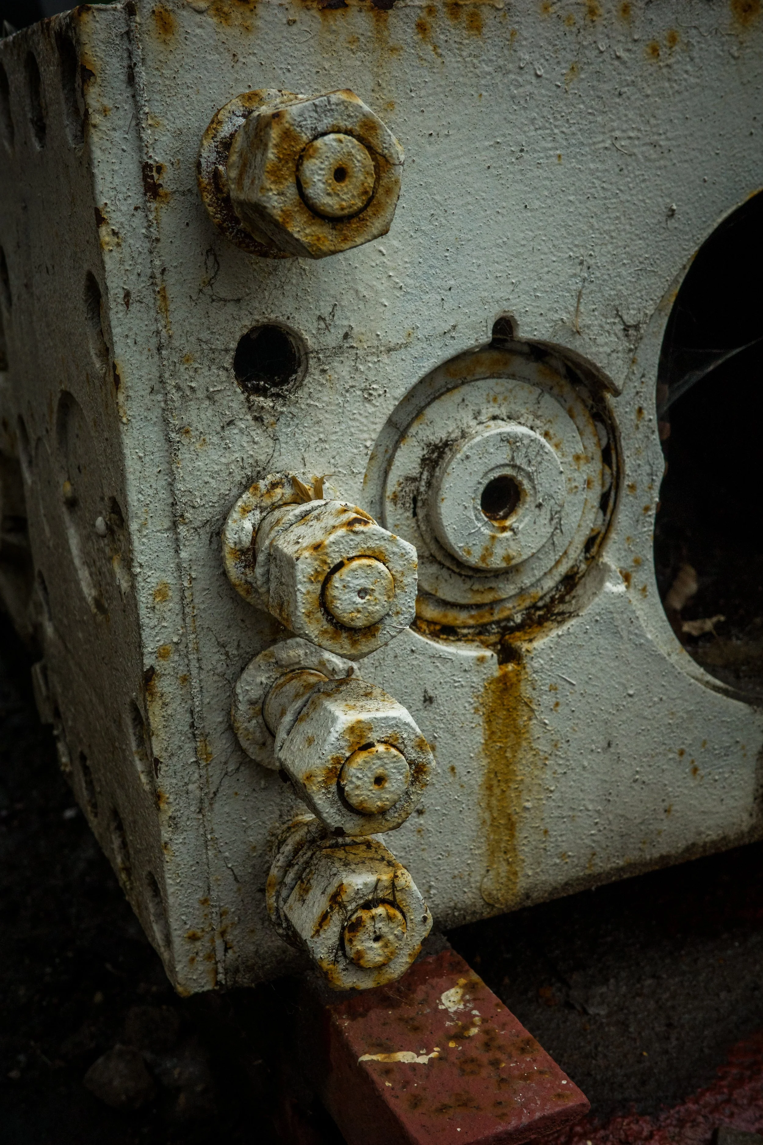 Close-up of a rusty, weathered industrial machine part with bolts and circular components, mounted on a textured, dirty concrete wall with a piece of red brick underneath.