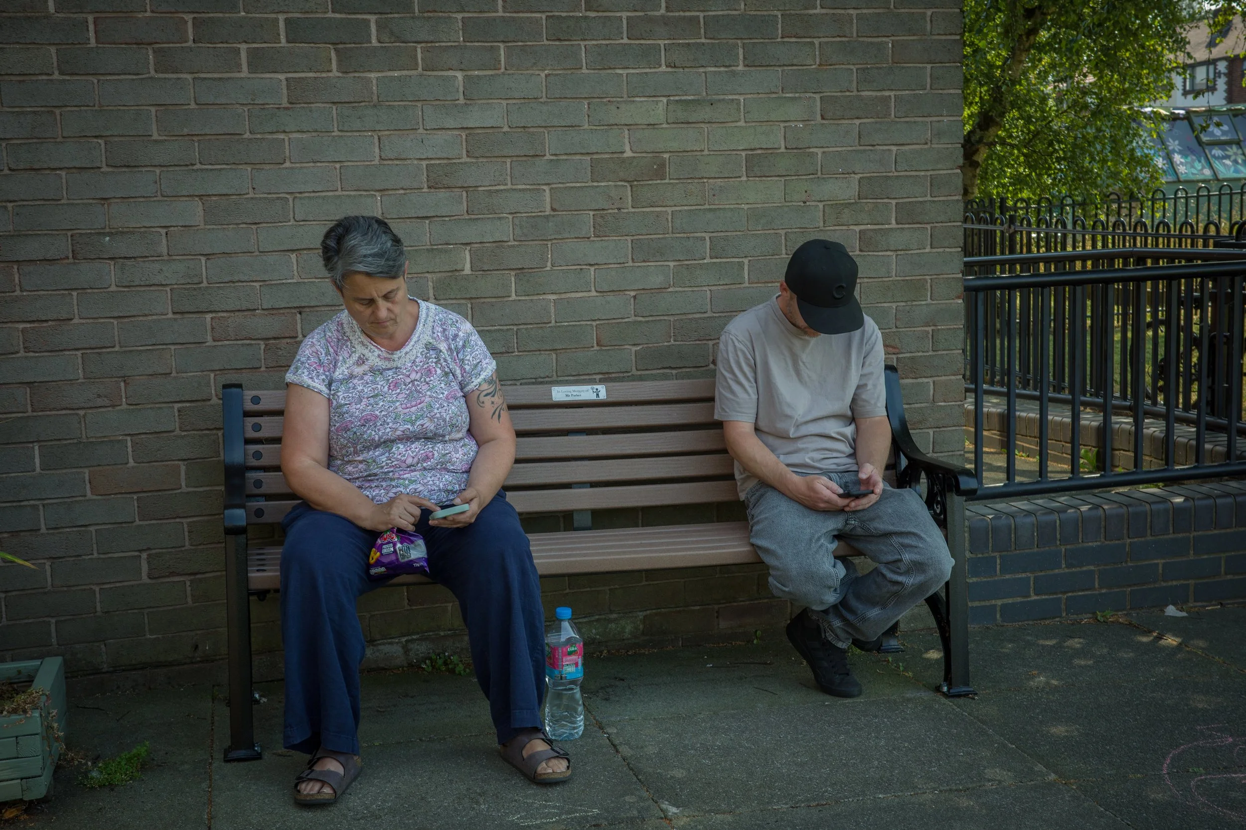 An elderly woman and man sitting on a bench outside, both looking at their phones. The woman has short gray hair and wears a floral shirt, while the man wears a black cap and a light gray t-shirt. There are trees and a brick wall in the background.