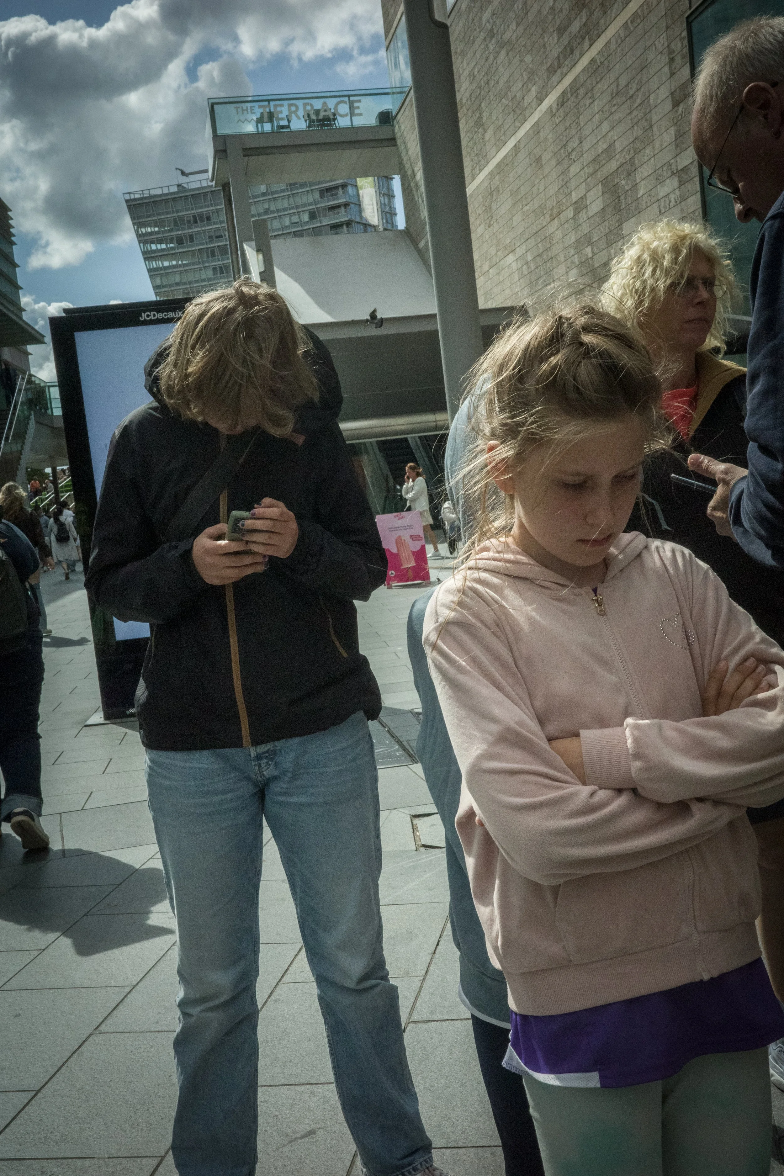 A group of people standing outside in a busy urban area, with a woman looking down at her phone, a young girl with arms crossed, and others engaged in conversation, under a cloudy sky.