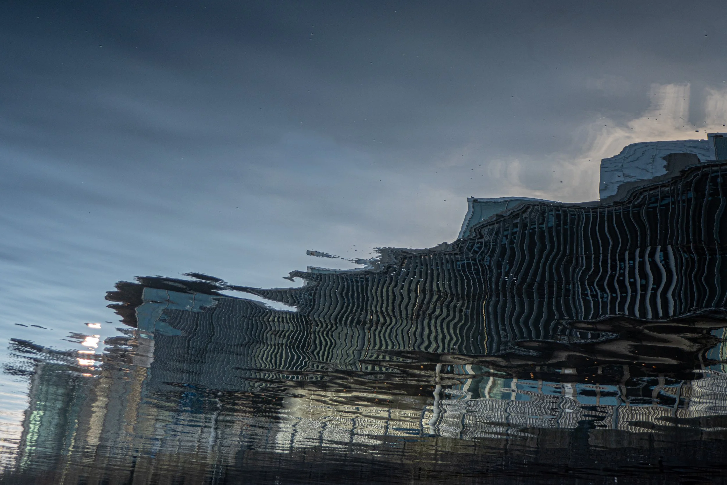Reflection of modern high-rise buildings on water with ripples, under cloudy sky.
