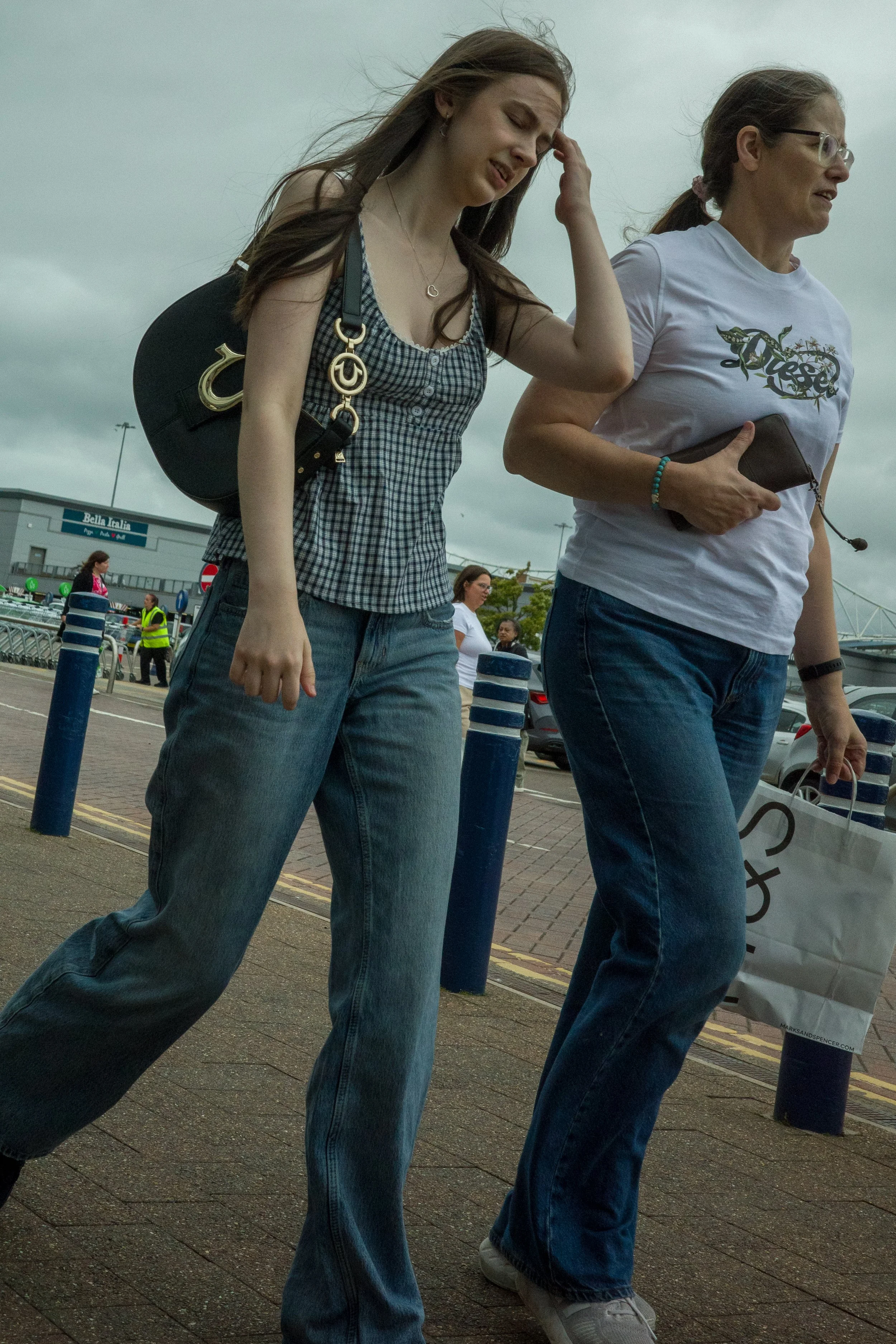Two women walking outdoors on a cloudy day at a parking lot, one carrying a black handbag and the other holding a shopping bag.