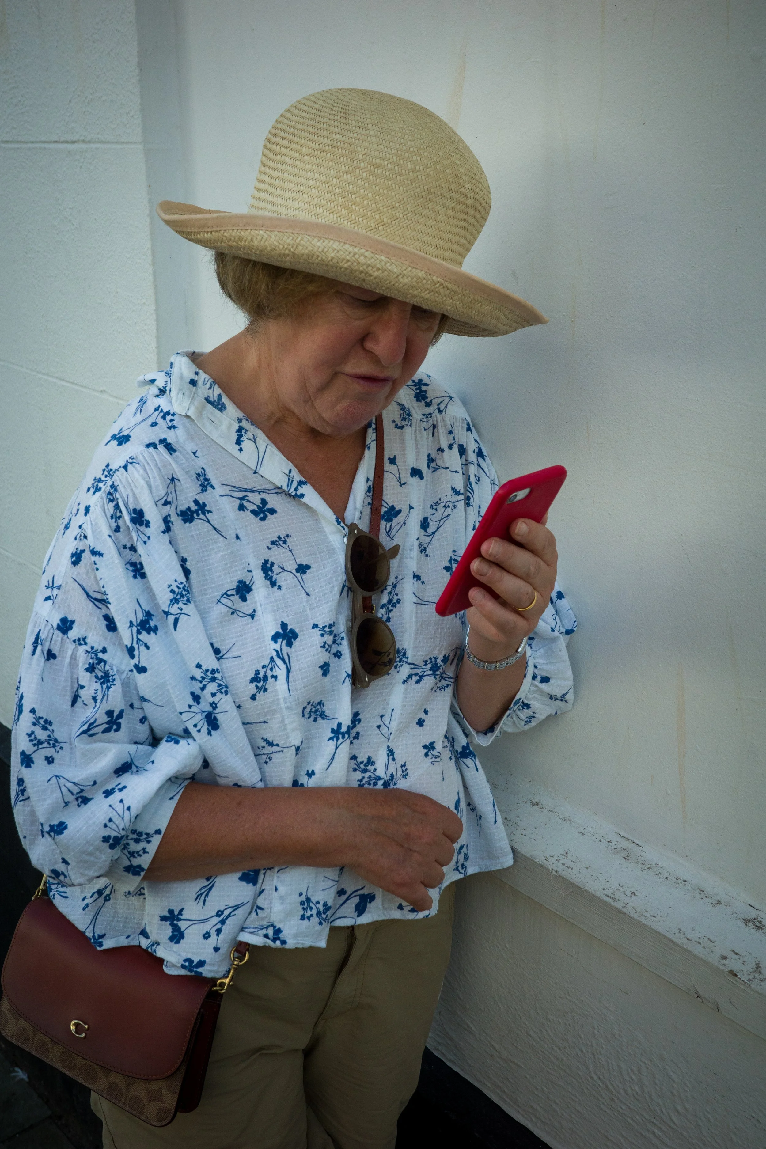An elderly woman with short, light-colored hair wearing a broad straw hat, a white blouse with blue floral pattern, khaki pants, and sunglasses hanging from her collar, standing beside a white wall while looking at her red smartphone.