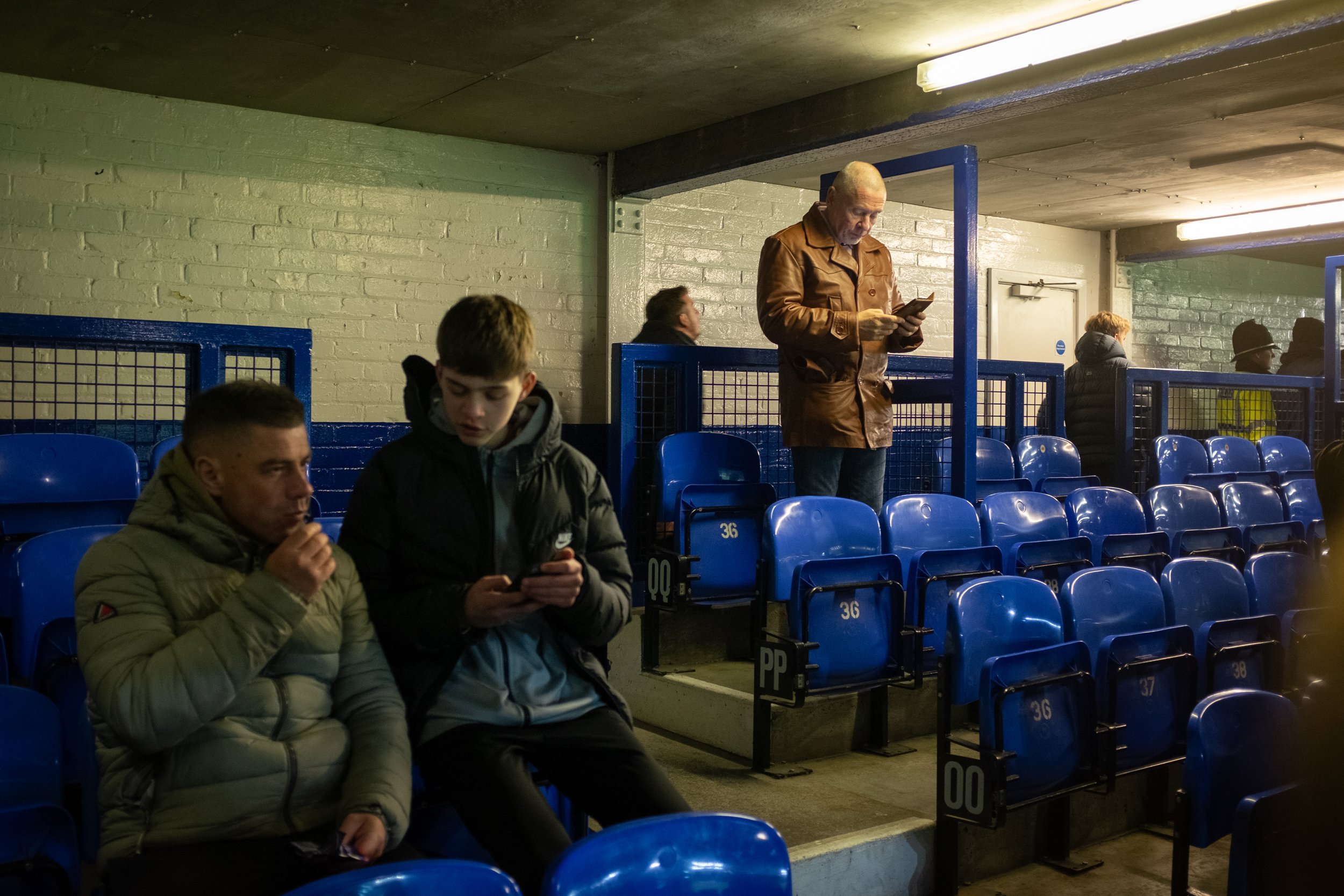 A group of people sitting in empty blue stadium seats with a white brick wall and ceiling in the background. Four individuals are visible, two in the foreground looking at their phones, one standing reading a phone, and another standing and talking w