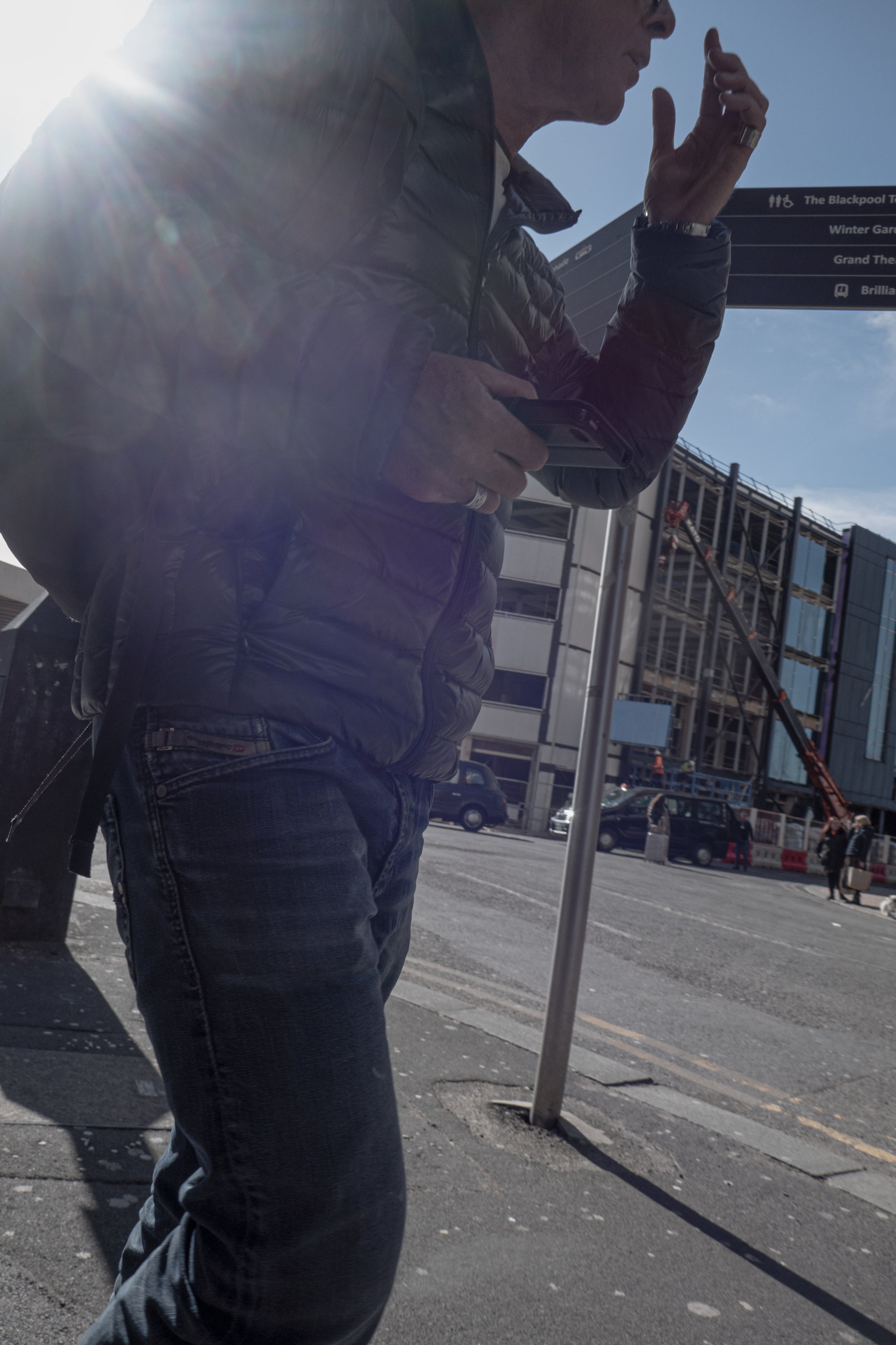 A man standing outdoors in sunlight, holding a phone, with a building and a signpost in the background.