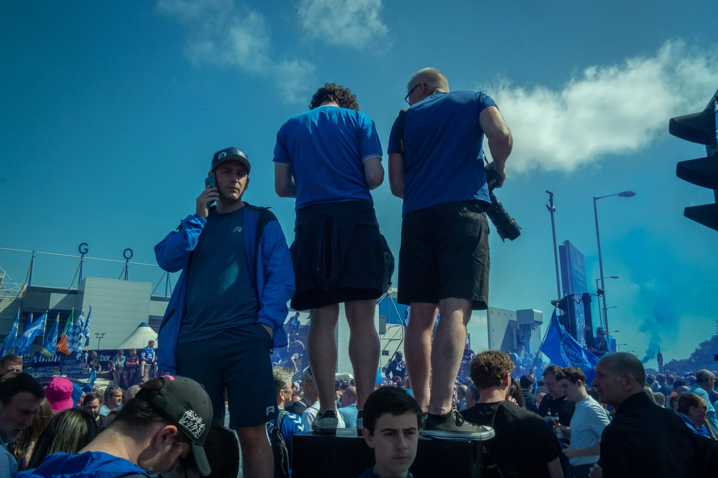 Crowd of people at a large outdoor event, with three individuals standing on a platform, one talking on a phone, others looking around, and many others in the background. The sky is mostly cloudy with some blue showing.