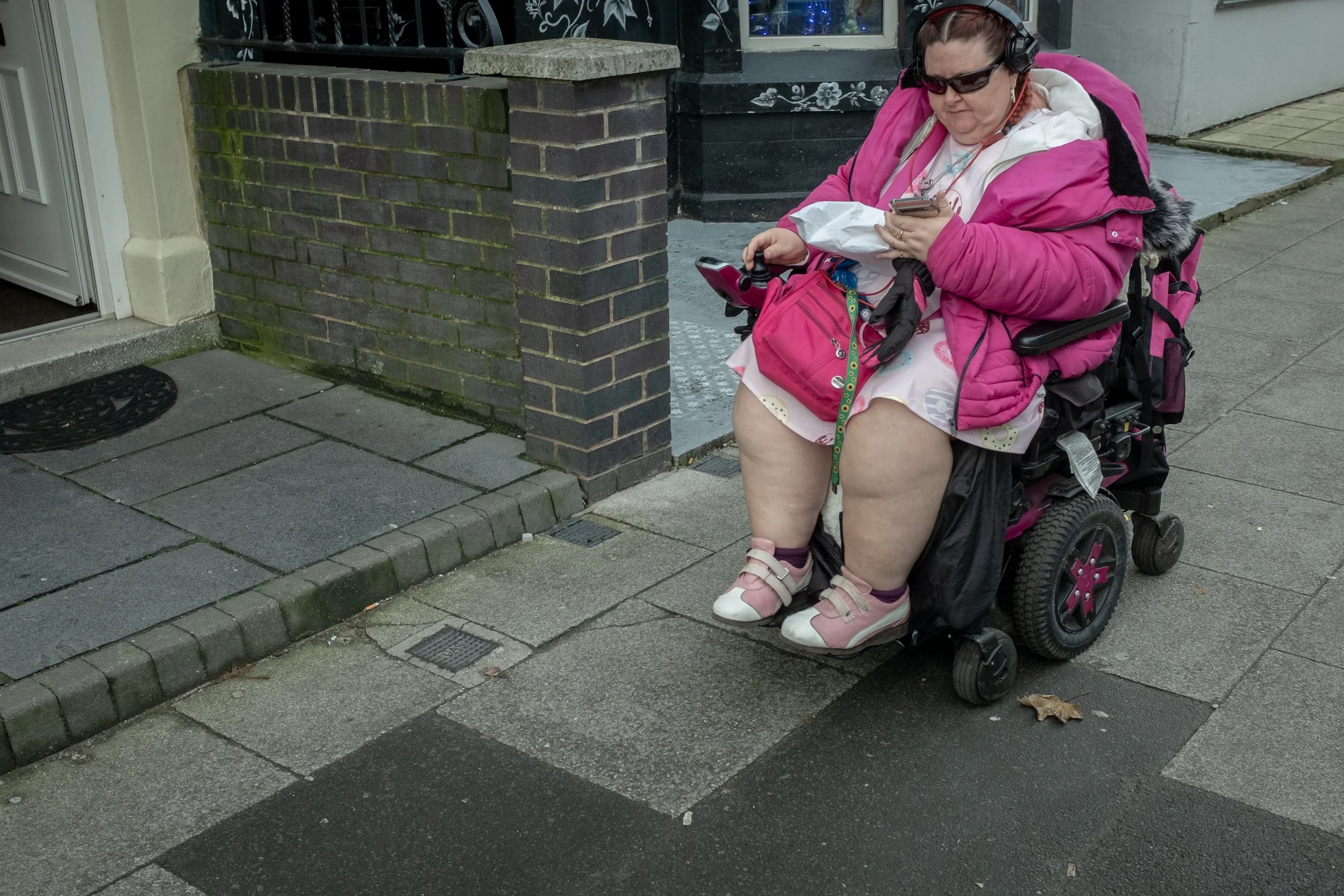 A woman with disabilities sitting in a motorized wheelchair on a sidewalk, wearing a pink jacket and white dress, looking at her phone, with a bag on her lap and headphones on her head.