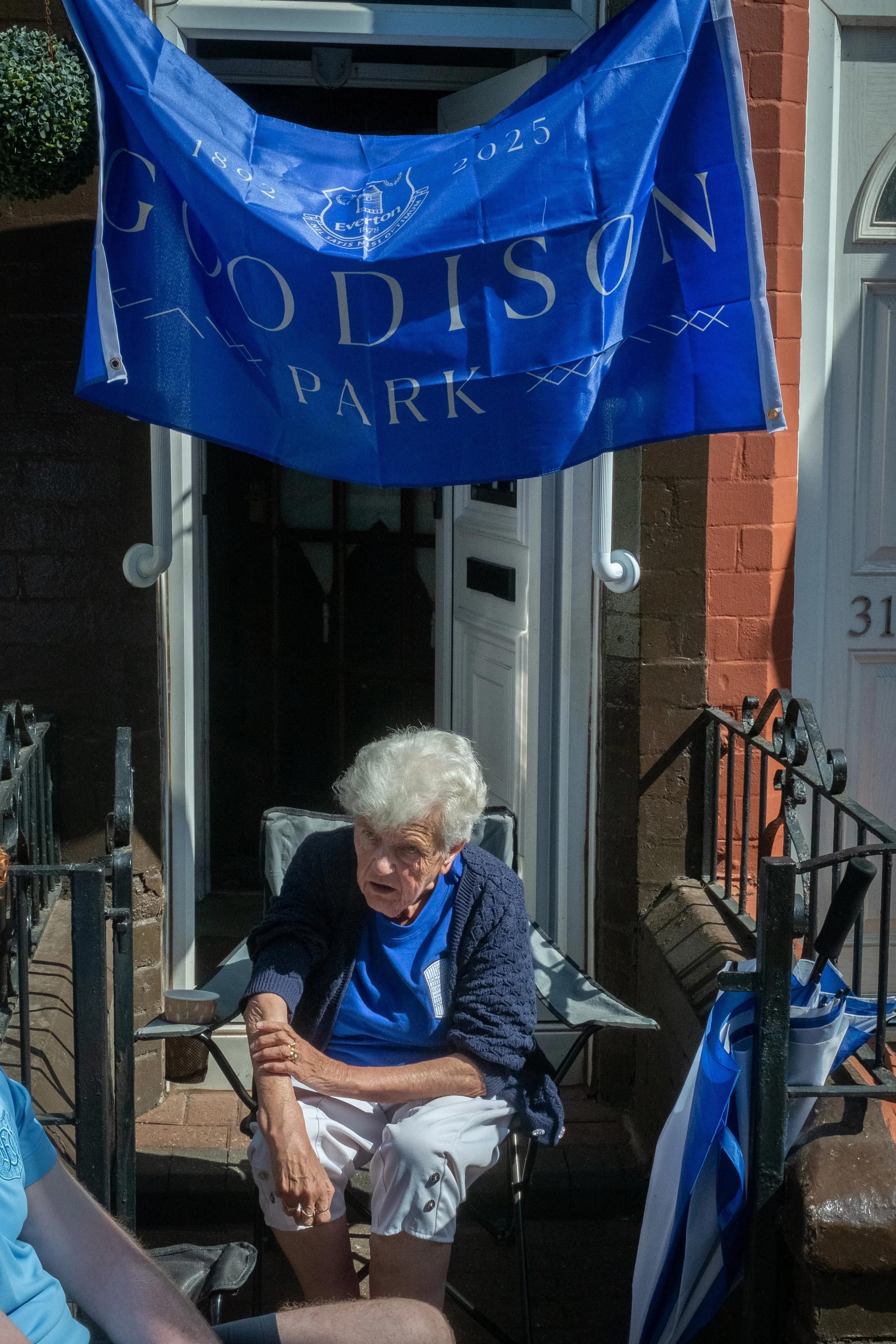 An elderly woman sitting outside a house, wearing a blue shirt and a dark sweater, with a Hummel’s Great Britain flag hanging above her. She appears to be in a festive setting for the Everton football team celebration.