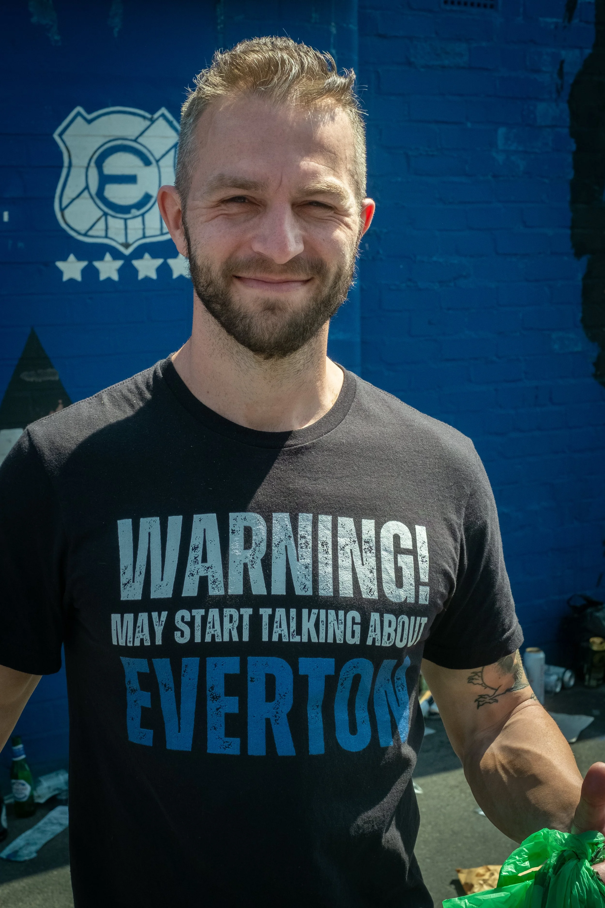 A man smiling outdoors in front of a blue brick wall with graffiti. He is wearing a black T-shirt that says 'WARNING! MAY START TALKING ABOUT EVERTON' and has a tattoo on his left arm.