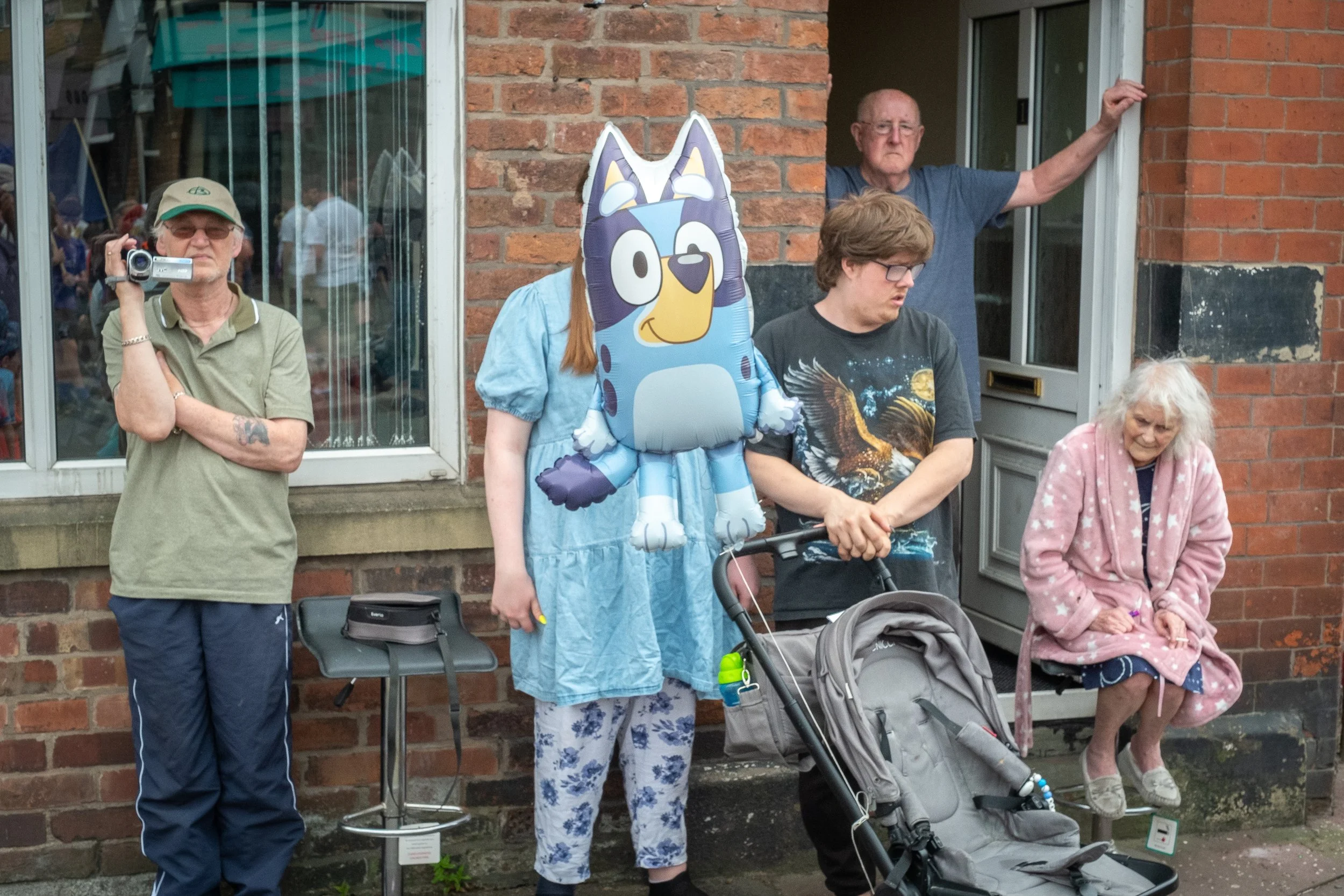 Group of people standing outside a brick building, including an elderly woman sitting on the window ledge wearing a pink robe, a young person with a wolf face balloon, an older man with glasses holding a camera, and several others observing or standi