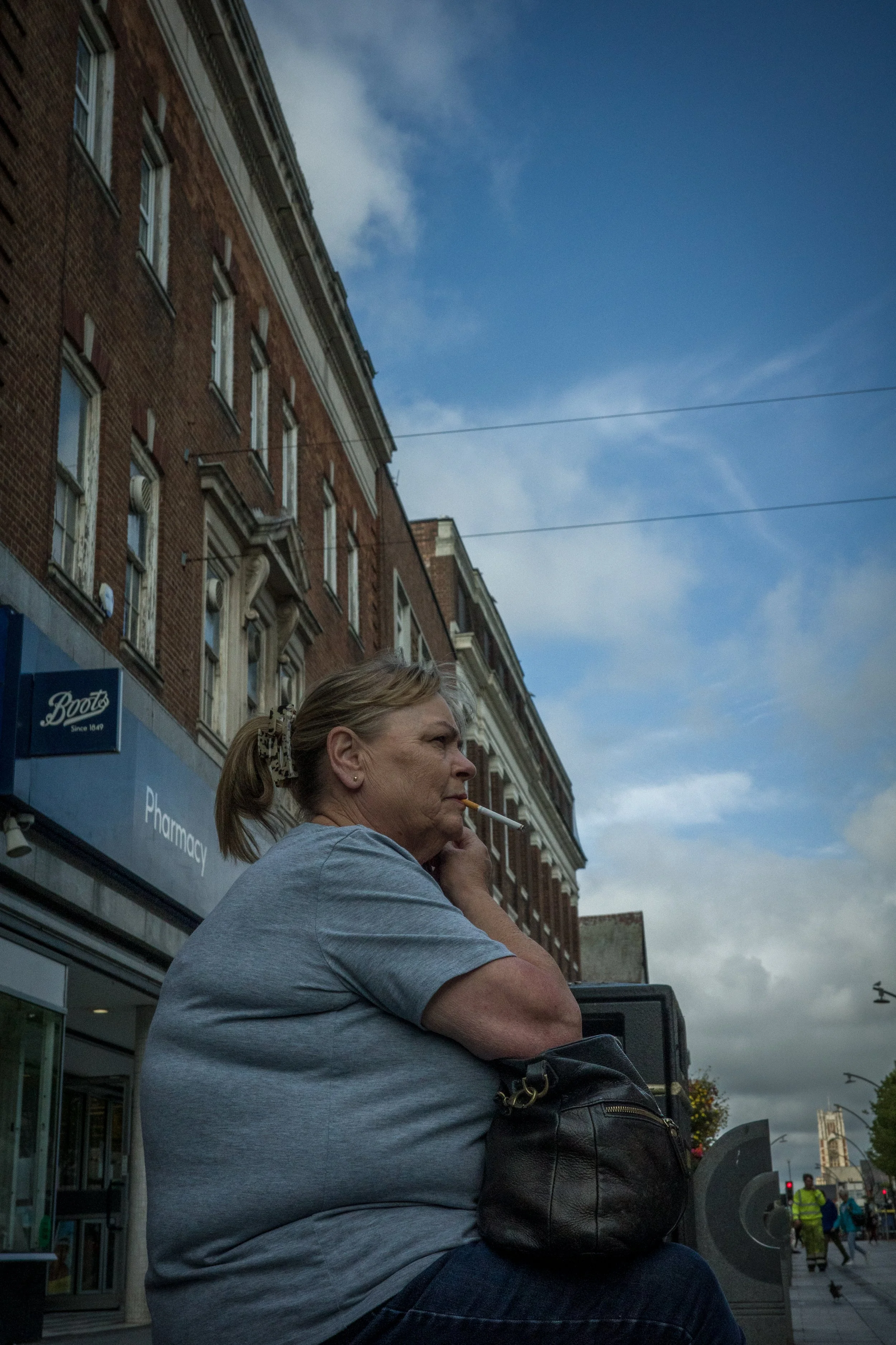 A woman sitting on a city street bench with a cigarette in her mouth, wearing a gray T-shirt, with brick buildings and a blue sky with clouds in the background.