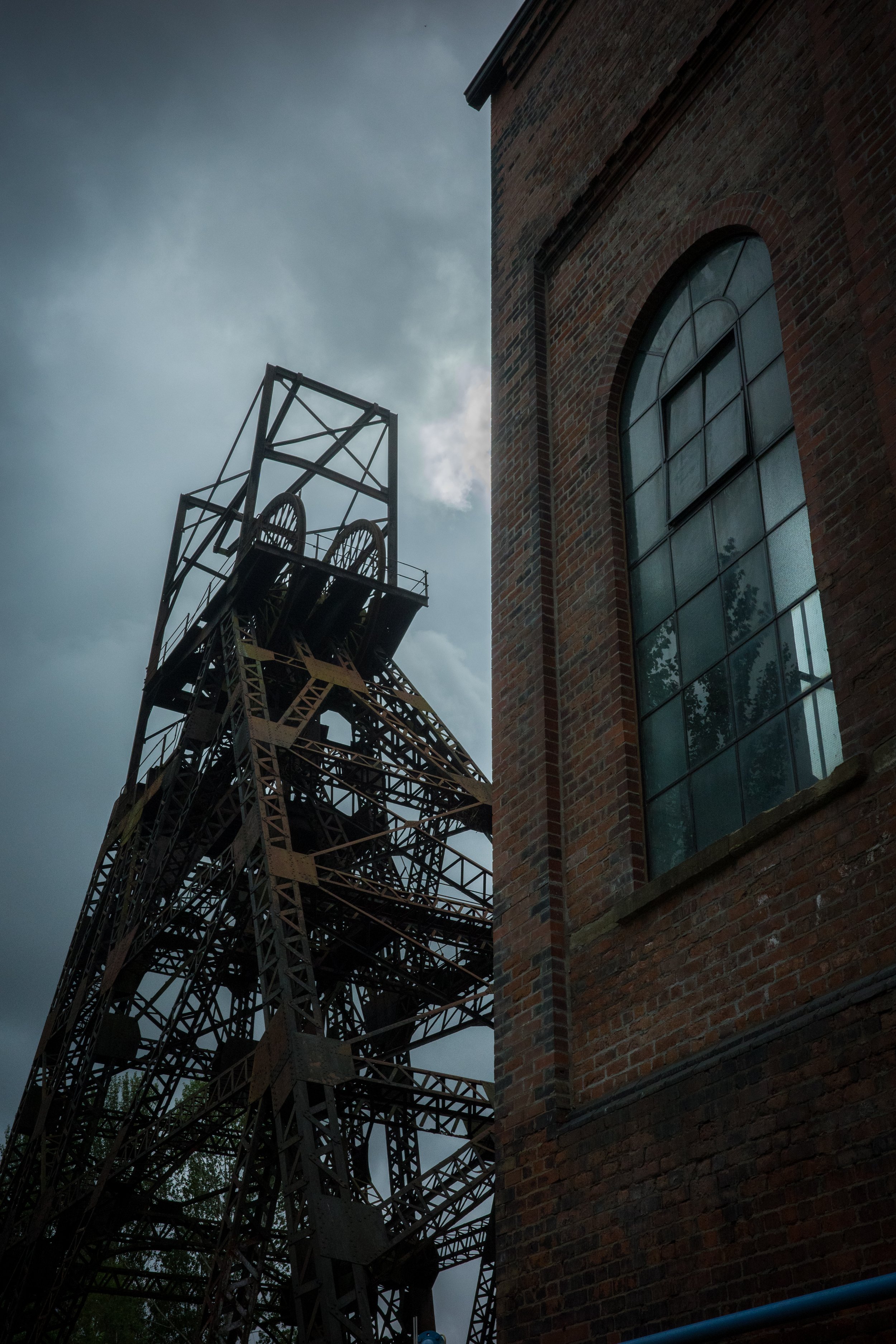 A tall metal industrial structure next to a brick building with a large arched window, under a cloudy sky.