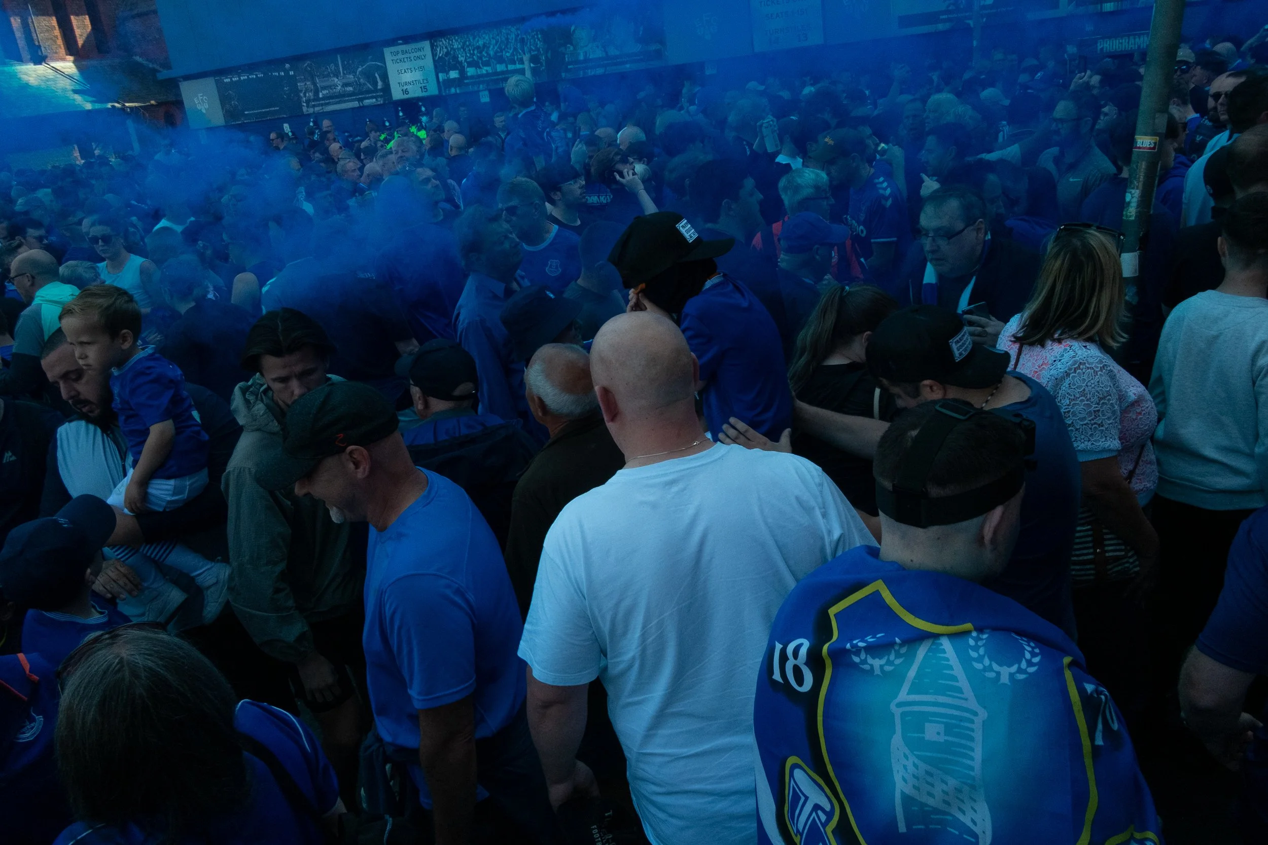 A large crowd of sports fans gathered outdoors, many wearing blue jerseys and team apparel, with some smoke or fog in the air.