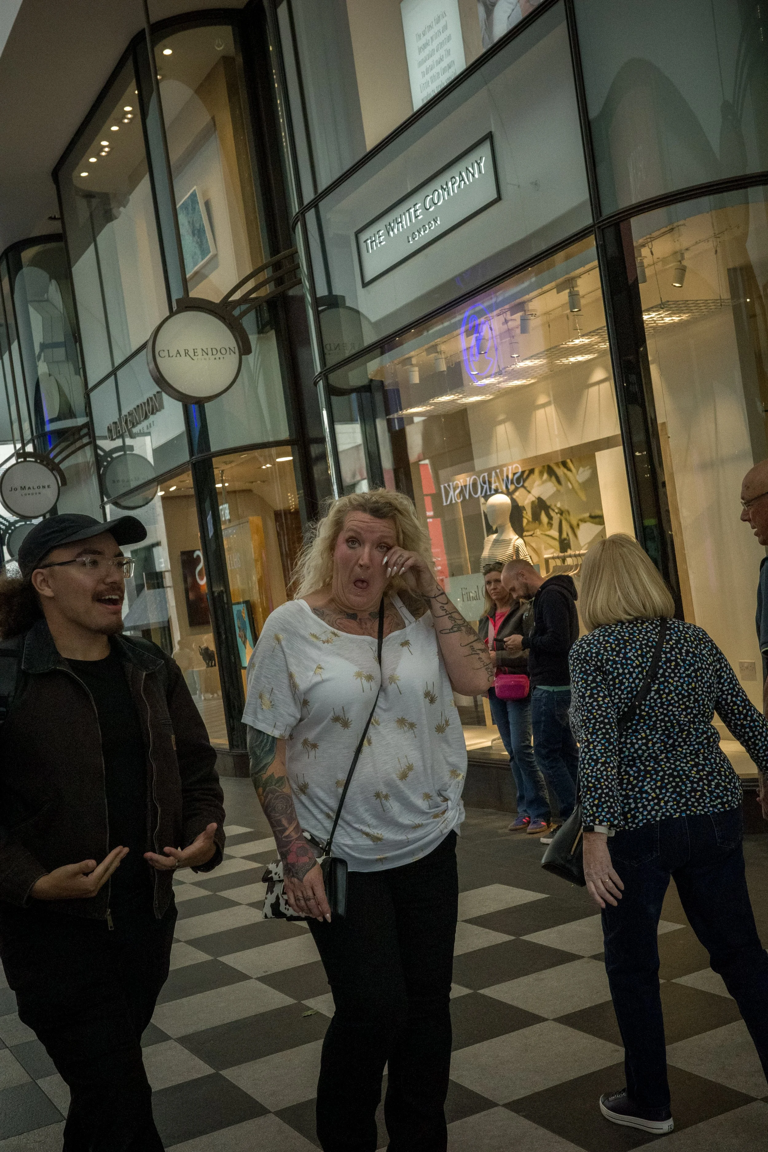 People walking in a shopping mall, with storefronts such as The White Company, Clarendon, and Jo Malone. One woman is wiping tears from her eyes, appearing emotional, while others are engaged in conversation around her.