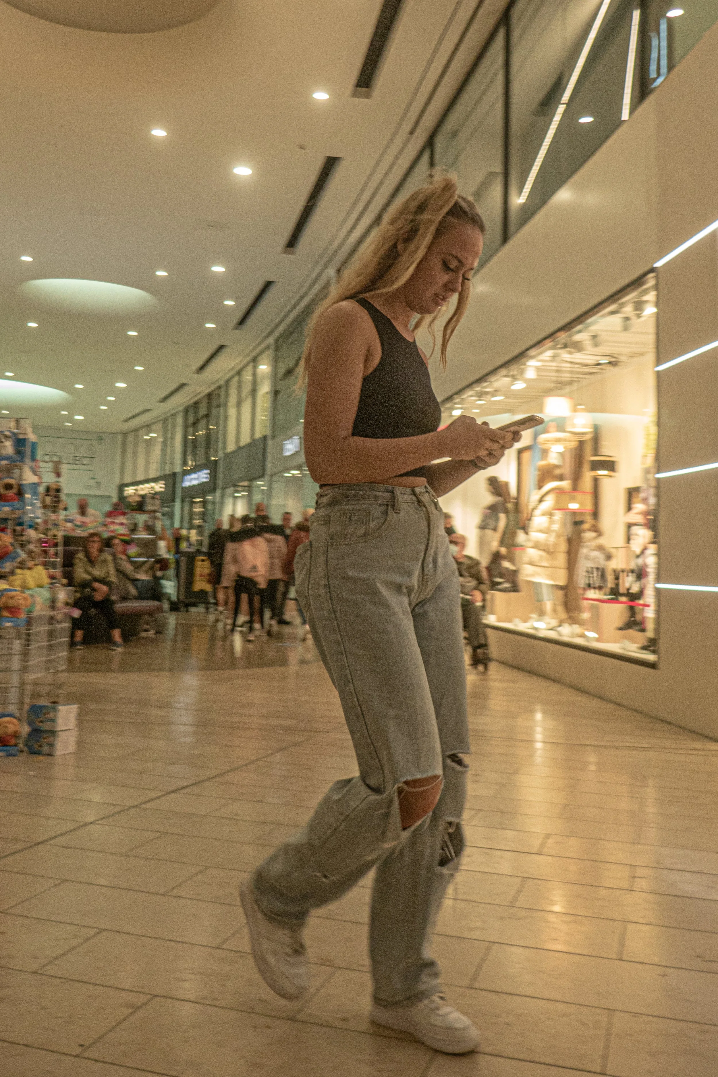 A young woman with blonde hair tied back is standing in a shopping mall, looking at her phone. She is wearing a black sleeveless top and ripped gray jeans with white sneakers. The mall has brightly lit stores in the background, and several people are