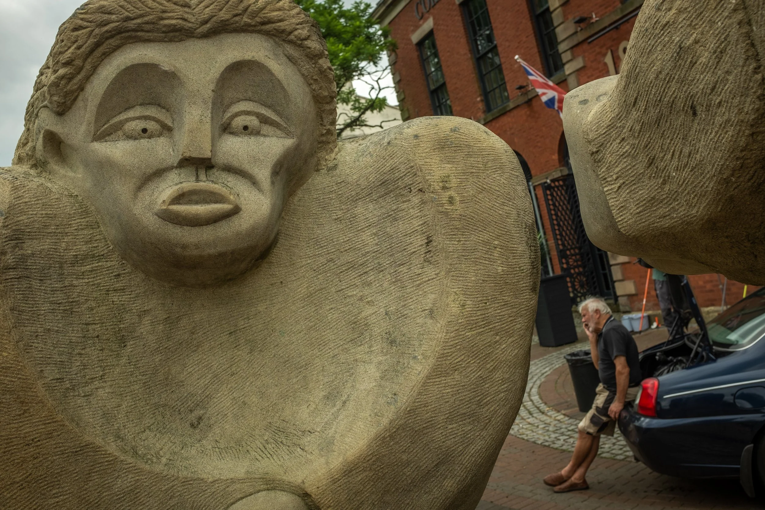 Close-up of a stone sculpture of a human face with detailed features, situated outdoors with a brick building, a person walking and a British flag in the background.