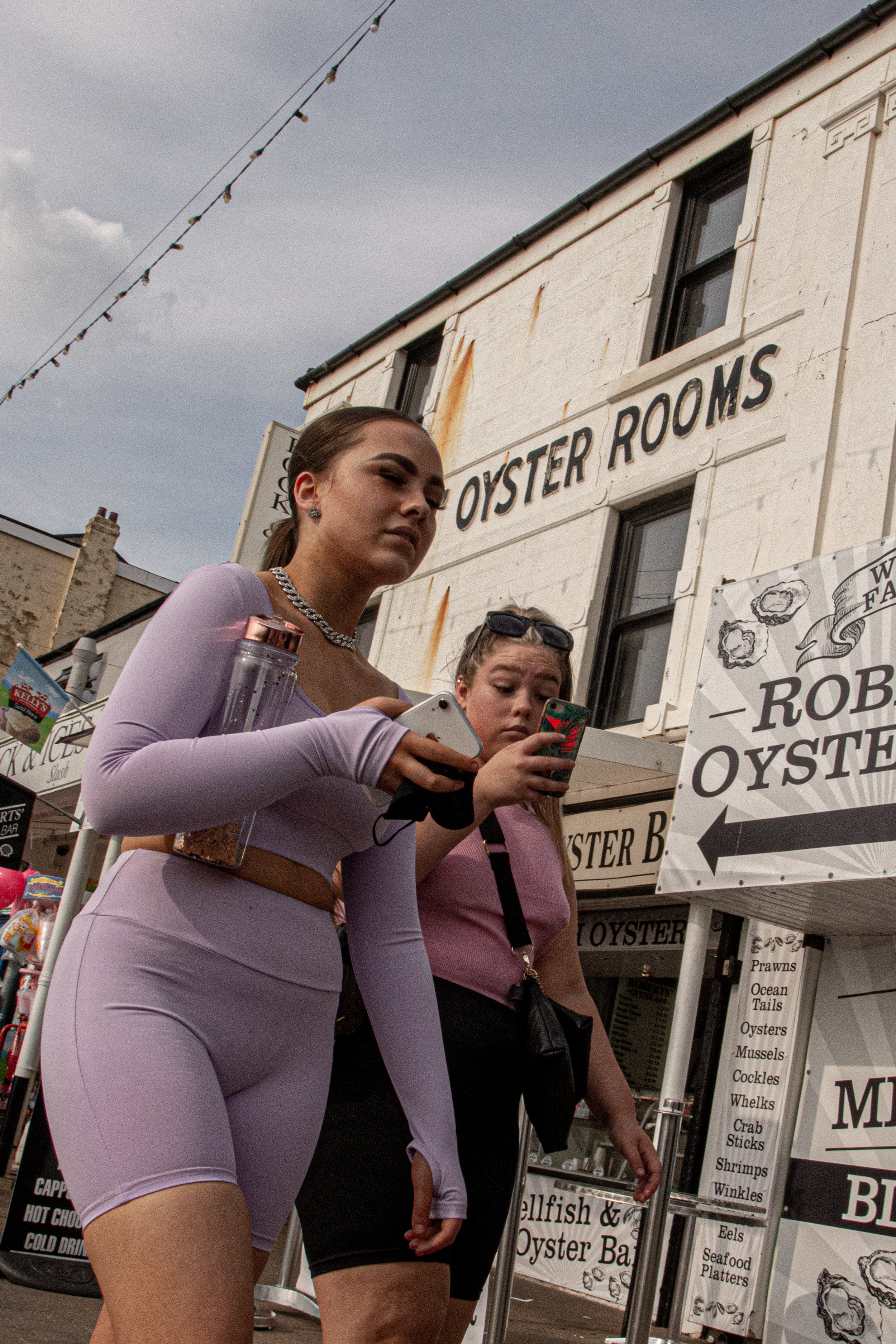 Two young women are standing near a food stand with signs for oysters and seafood in an outdoor market or fair setting. One woman is wearing a tight, lavender athletic outfit with a cutout, holding a phone in her hand, and has her hair pulled back. T