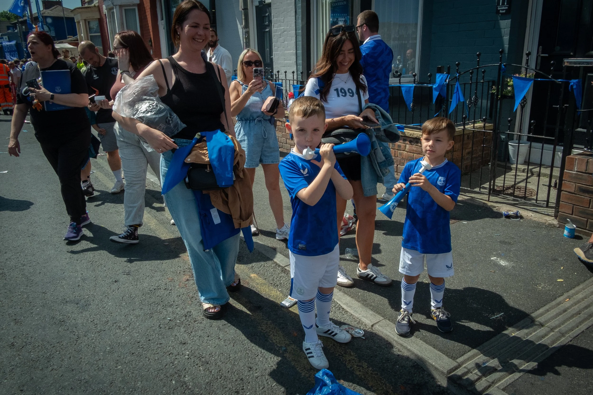 Two young boys in blue sports uniforms, playing trumpets, standing on a street with a group of smiling adults and other children around them at an outdoor event.