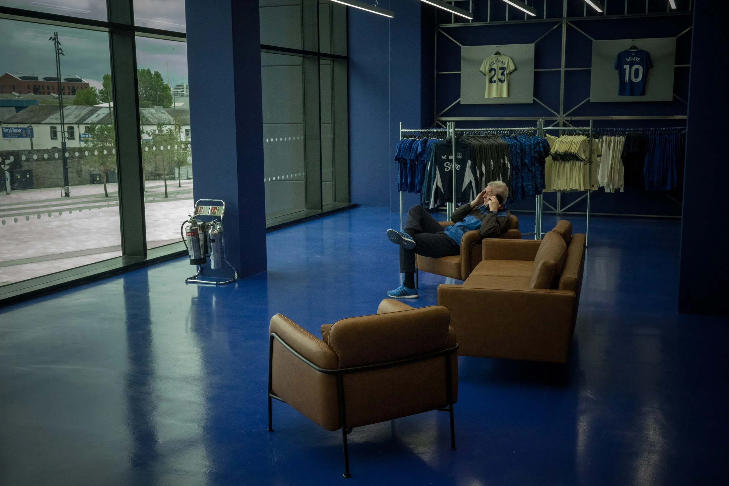 A man sitting on a beige couch talking on his cellphone inside a sports apparel store. The store has blue walls and displays soccer jerseys and shirts on racks and wall-mounted frames, including jerseys with the names Colema and Indaye, with the numb
