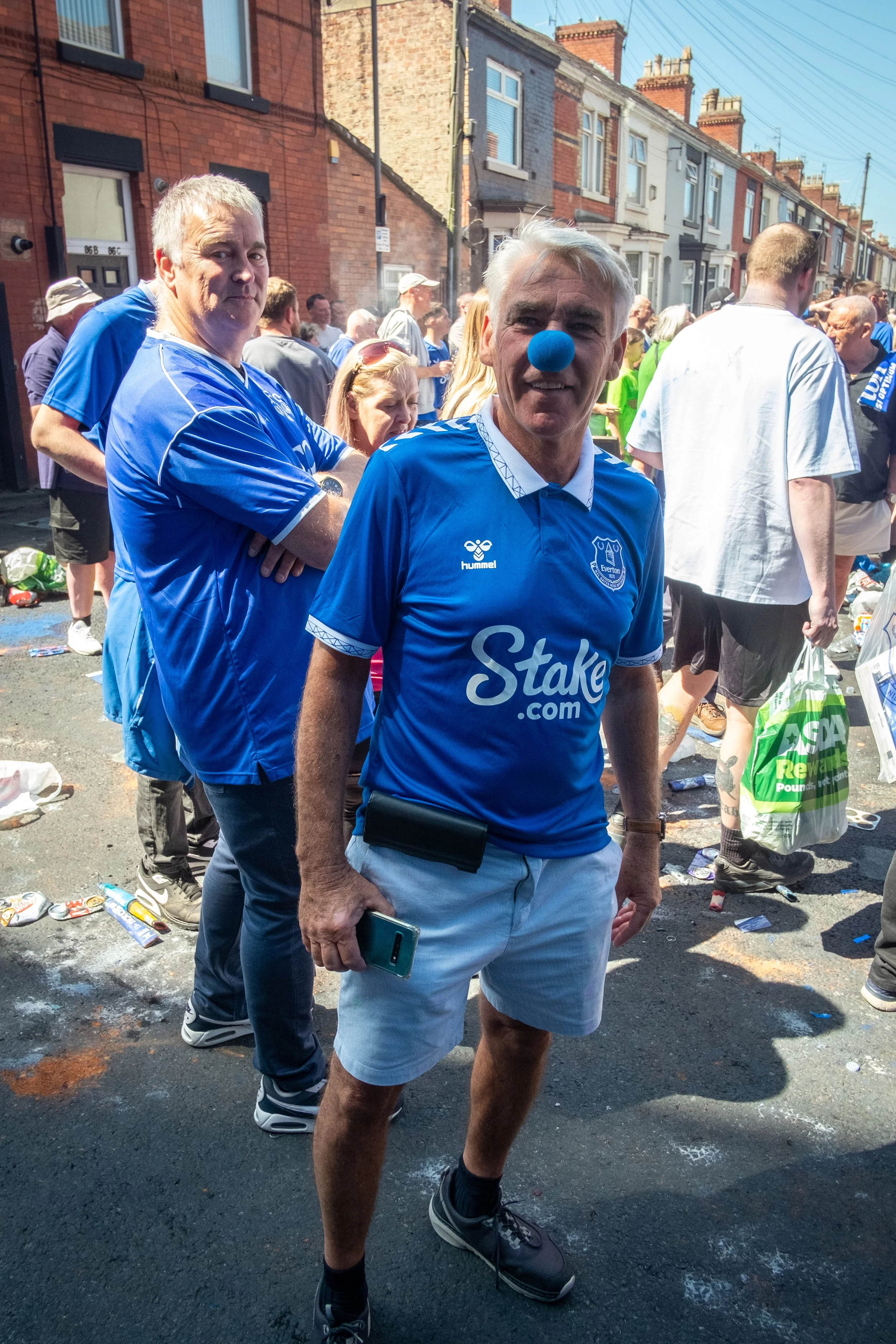 A man wearing a blue Everton jersey with a blue clown nose stands in a crowd on a city street, surrounded by people and debris.