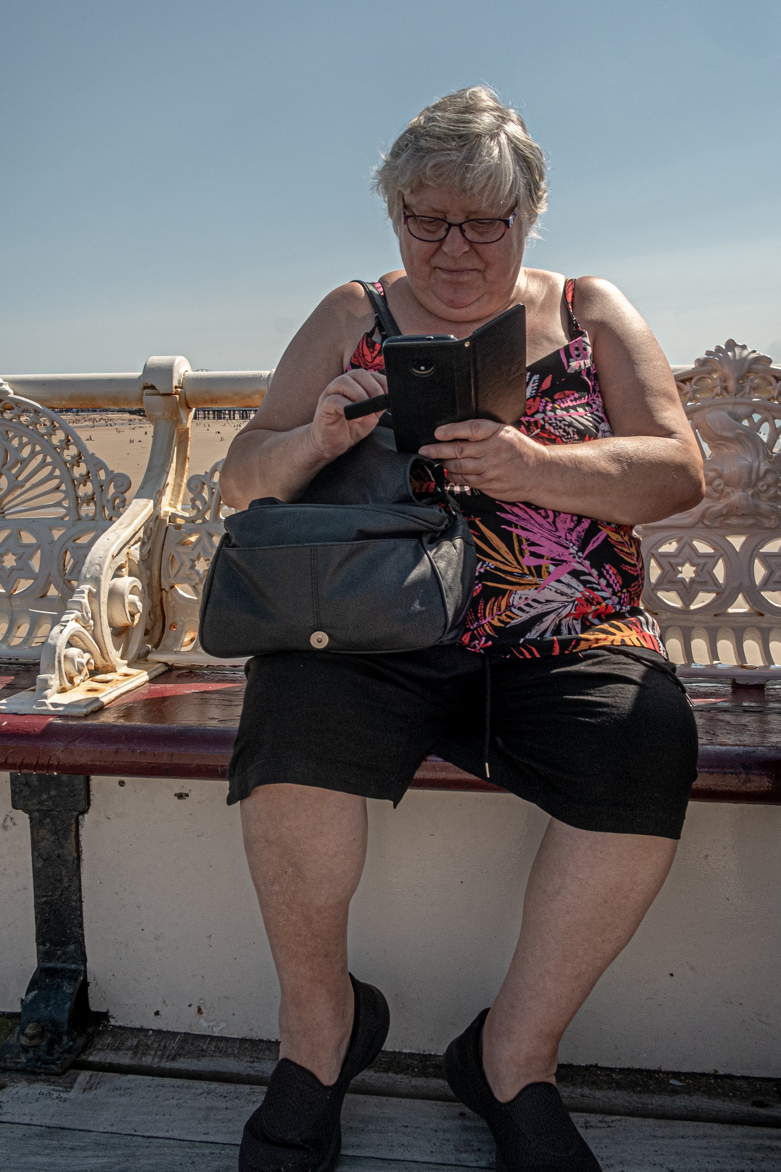 Older woman with glasses sitting on a decorative white metal bench at the beach, wearing a colorful sleeveless top and black shorts, looking at her phone.