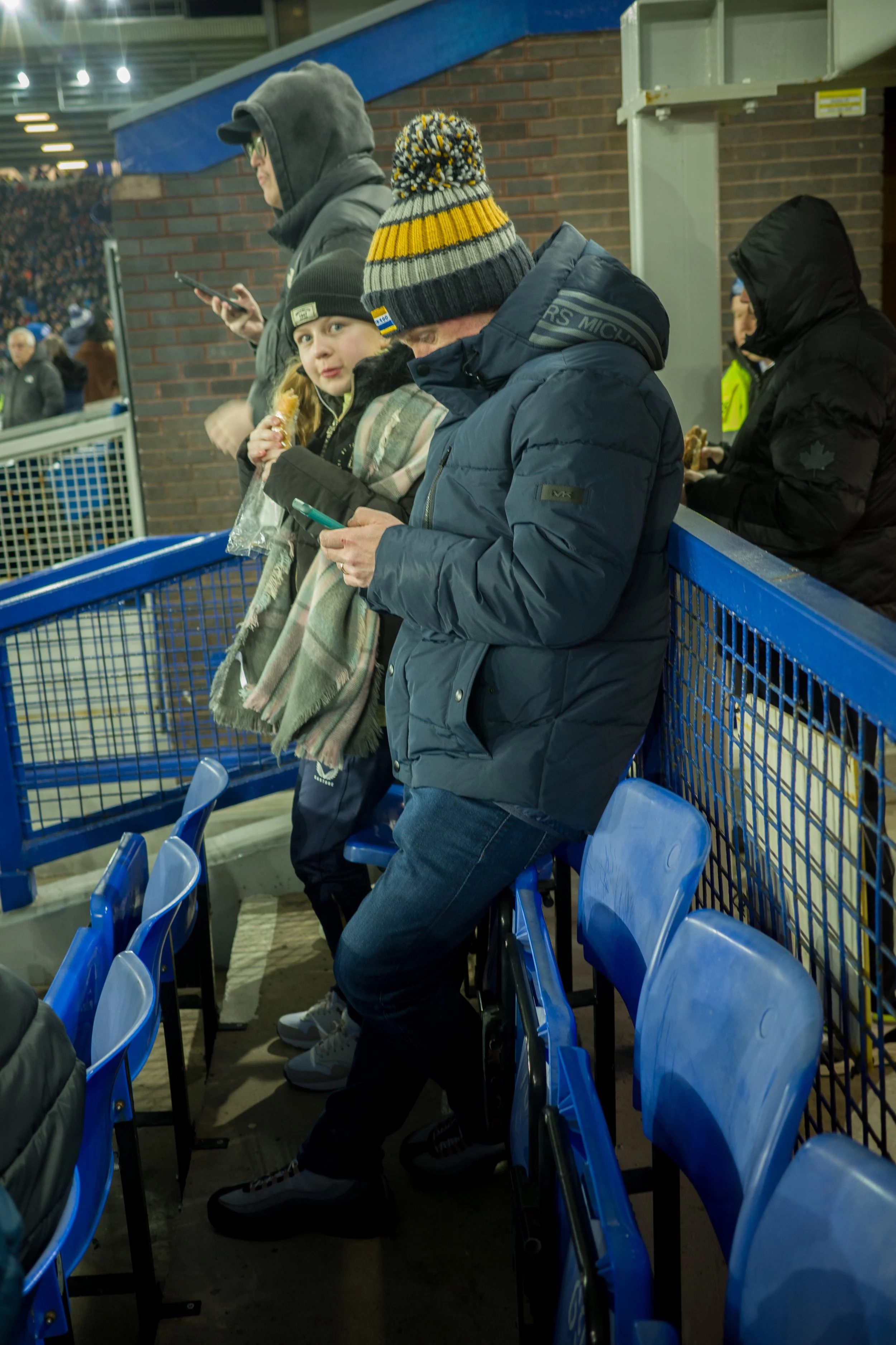People standing and sitting behind a blue fence at what appears to be a sports event, wearing winter clothing including hats and jackets.
