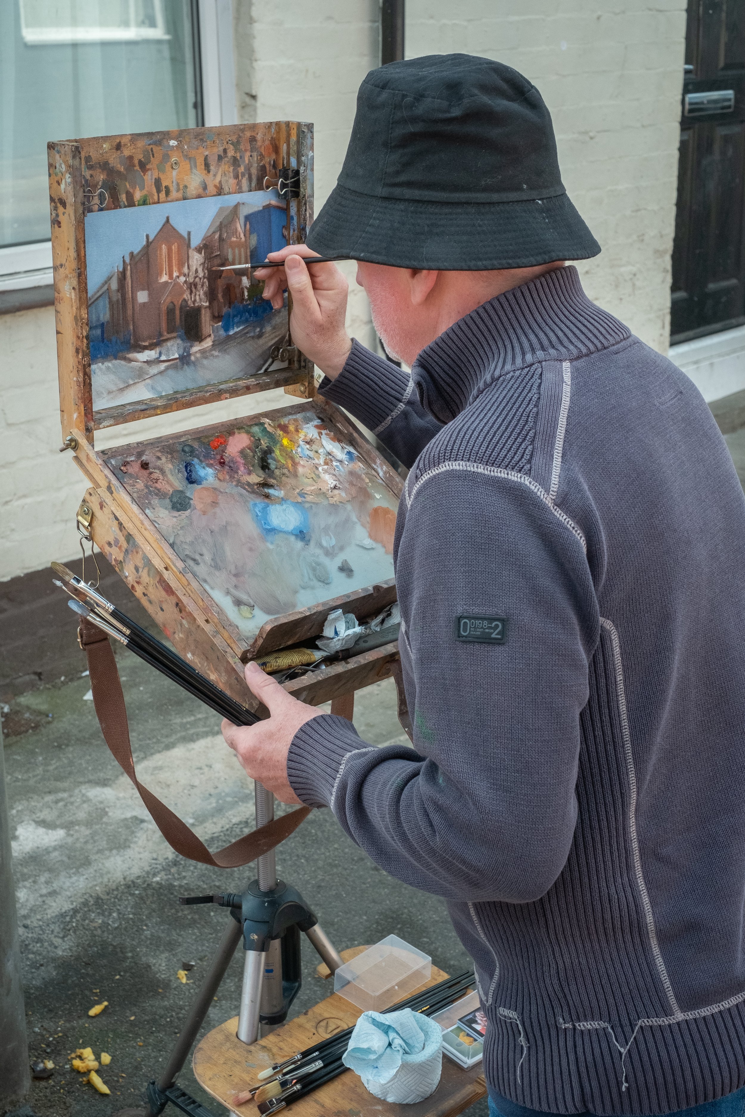 An elderly man painting a cityscape on an outdoor easel with a palette of mixed colors.