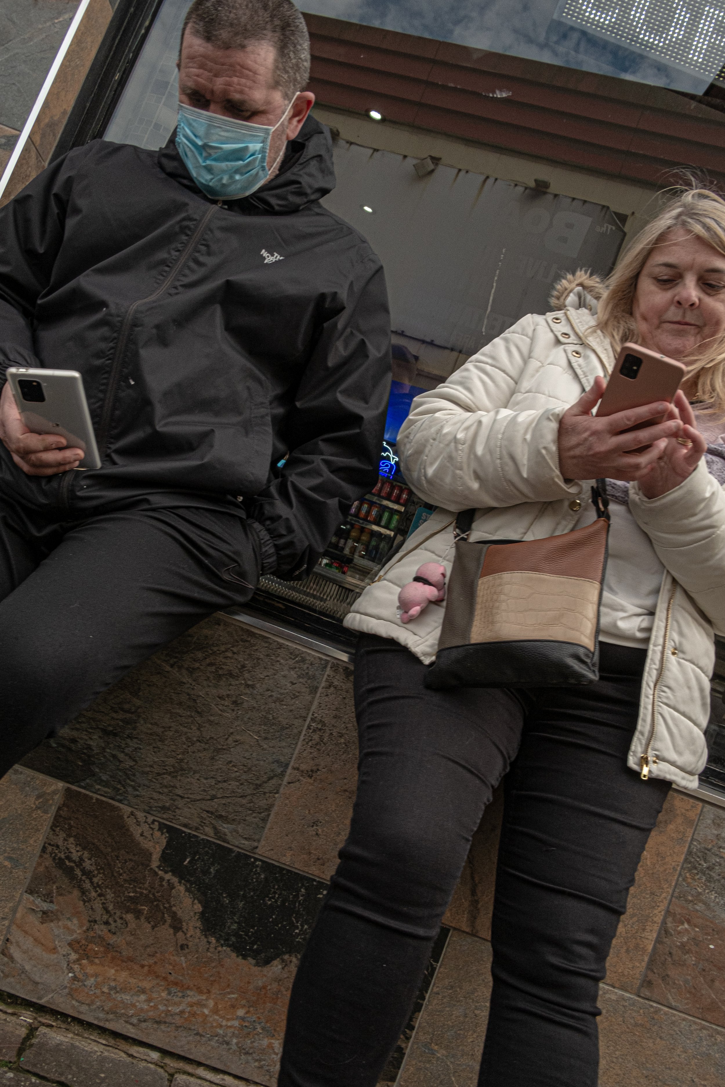 A man and a woman sitting on a bench, both looking at their phones. The man is wearing a black North Face jacket and a blue face mask, while the woman is wearing a white coat and glasses. They are outside a building with a glass window and a vending 