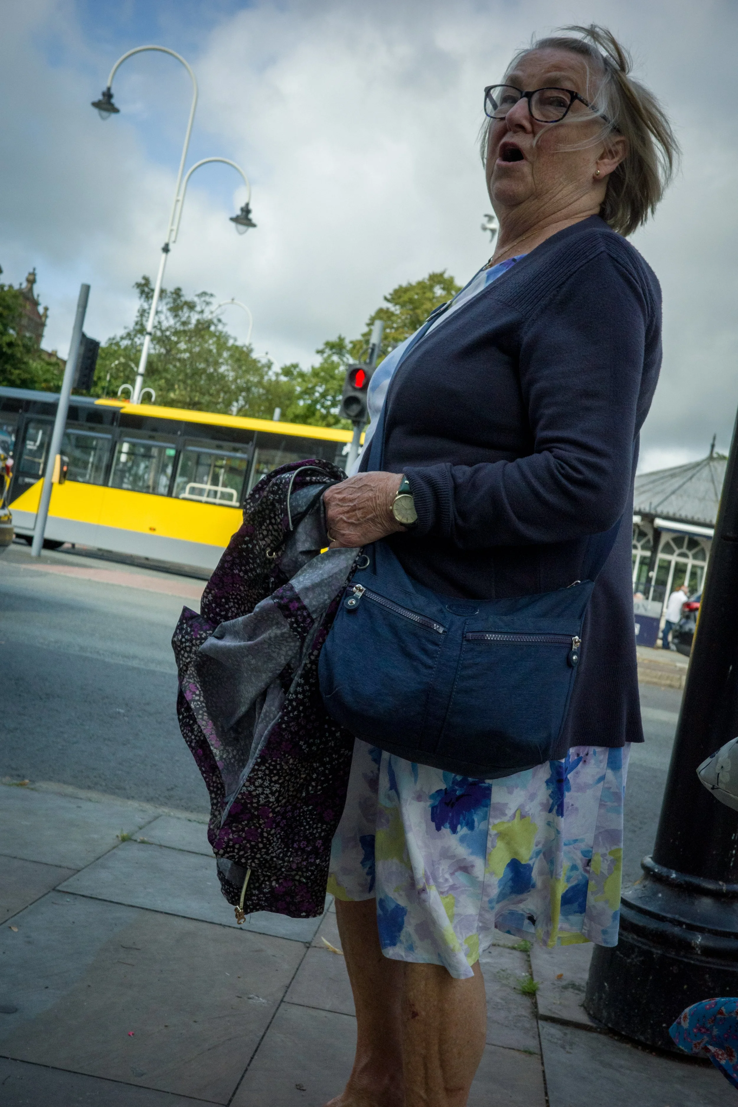 An elderly woman with gray hair, wearing glasses, a blue cardigan, and a floral skirt, stands on a city sidewalk near a black lamppost and a yellow tram with red traffic lights in the background.