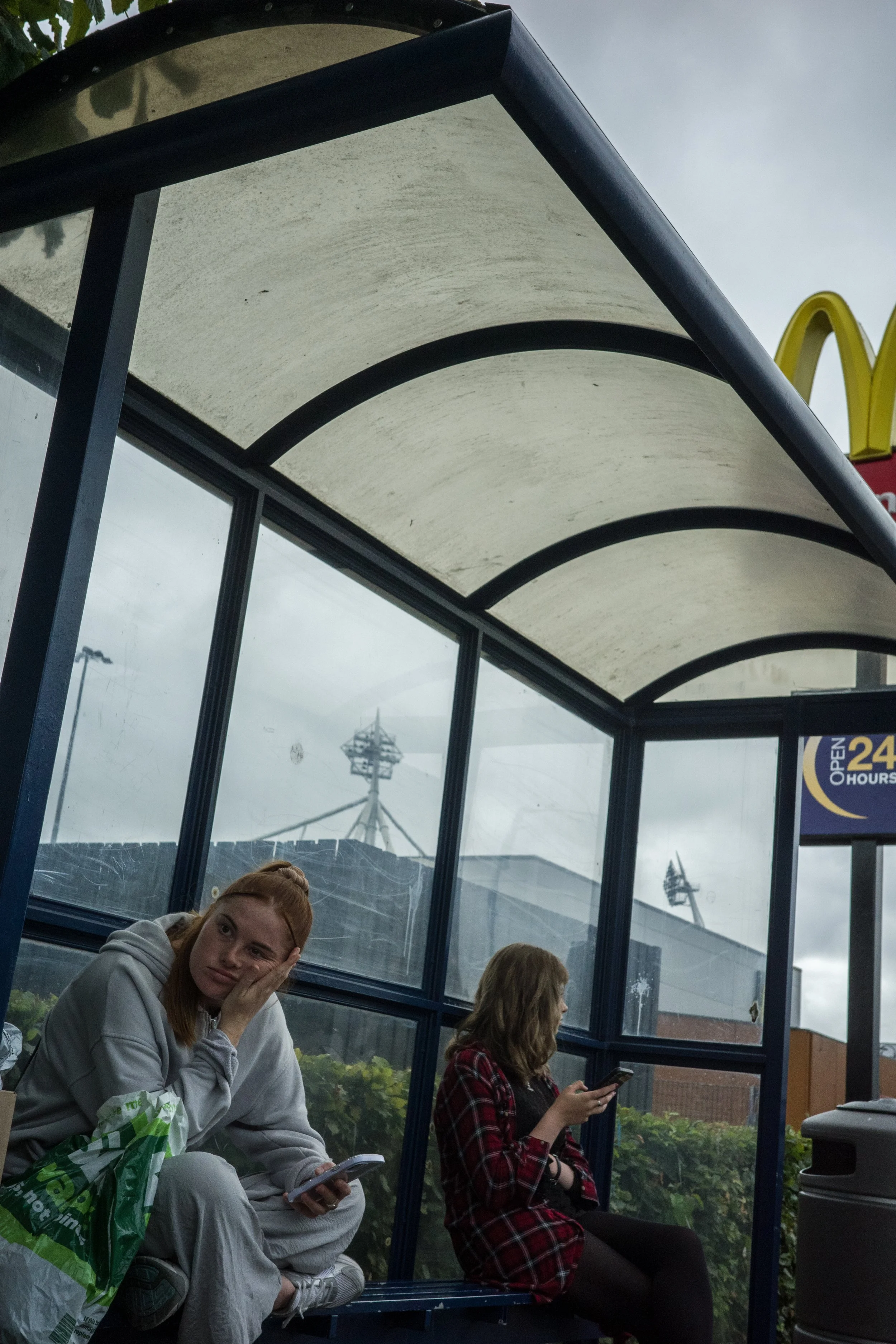 Two women sitting at a bus stop under a curved roof. One woman, with red hair and gray hoodie, is holding a phone and resting her head on her hand. The other woman, with brown hair and a red plaid shirt, is looking at her phone. Behind them, there ar