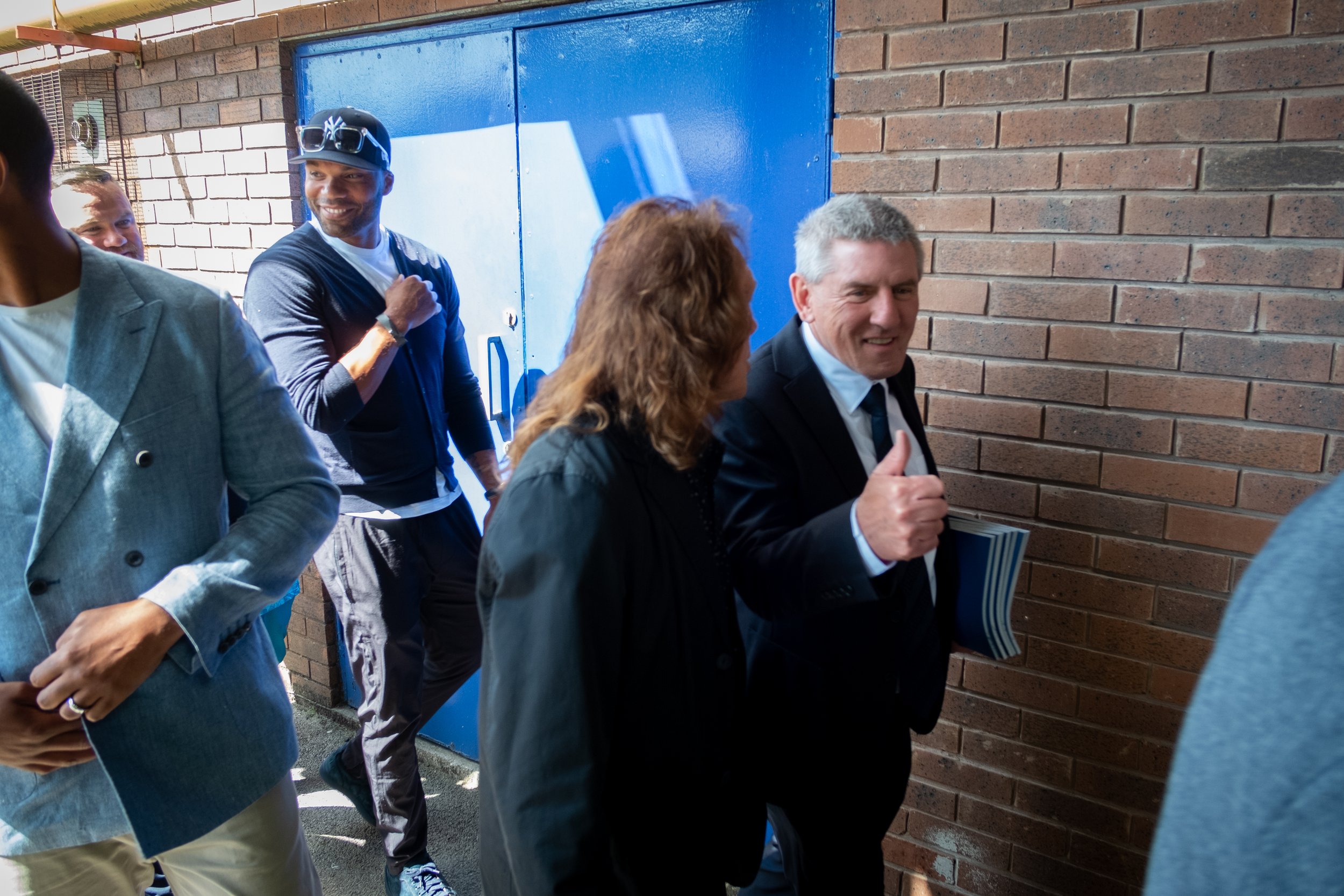 A group of people walking and talking outside, with a focus on a man in a suit giving a thumbs-up gesture.