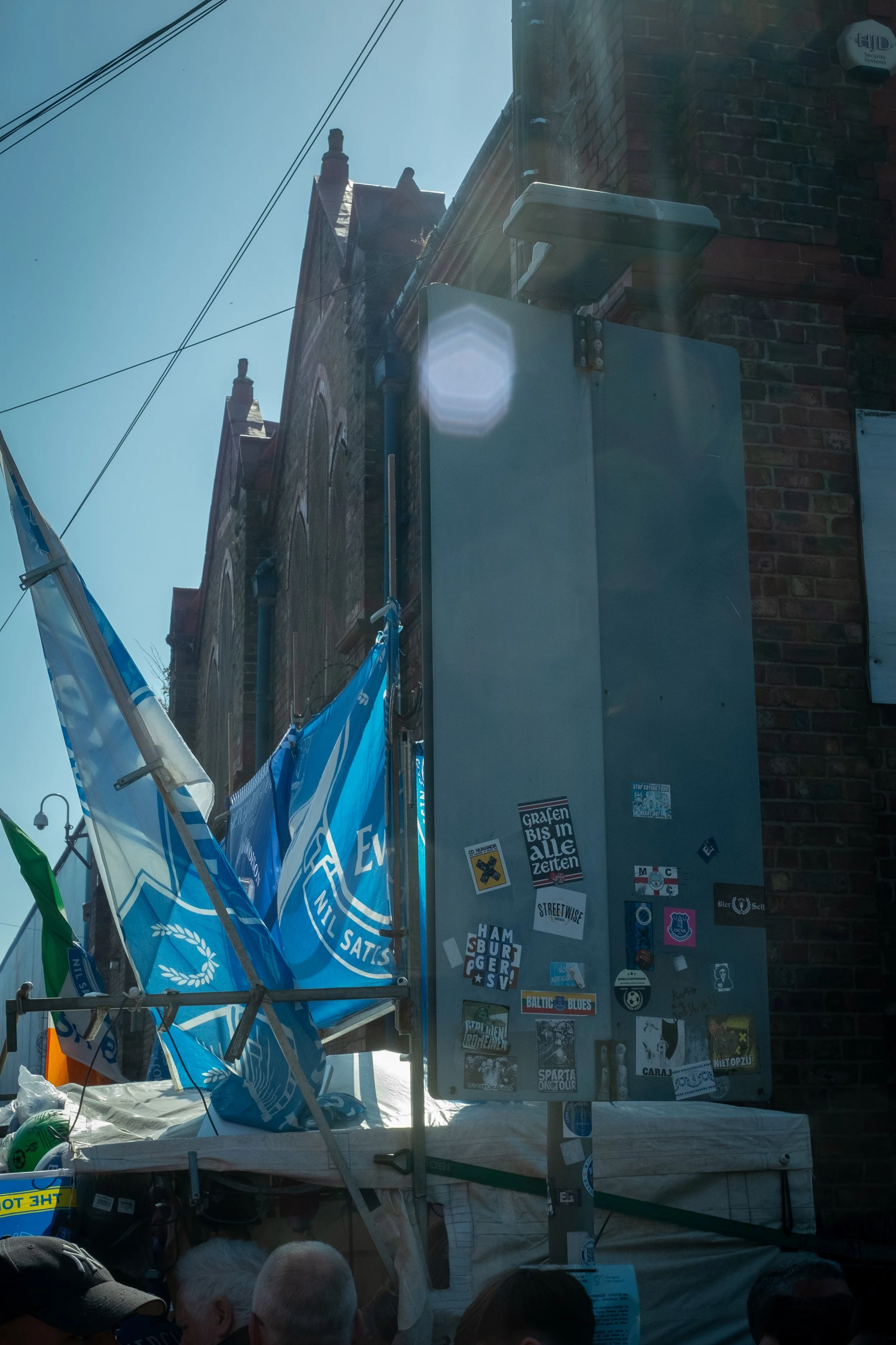 Street scene with blue flags, a brick building, and a pole covered with stickers. There are people partially visible at the bottom of the image.