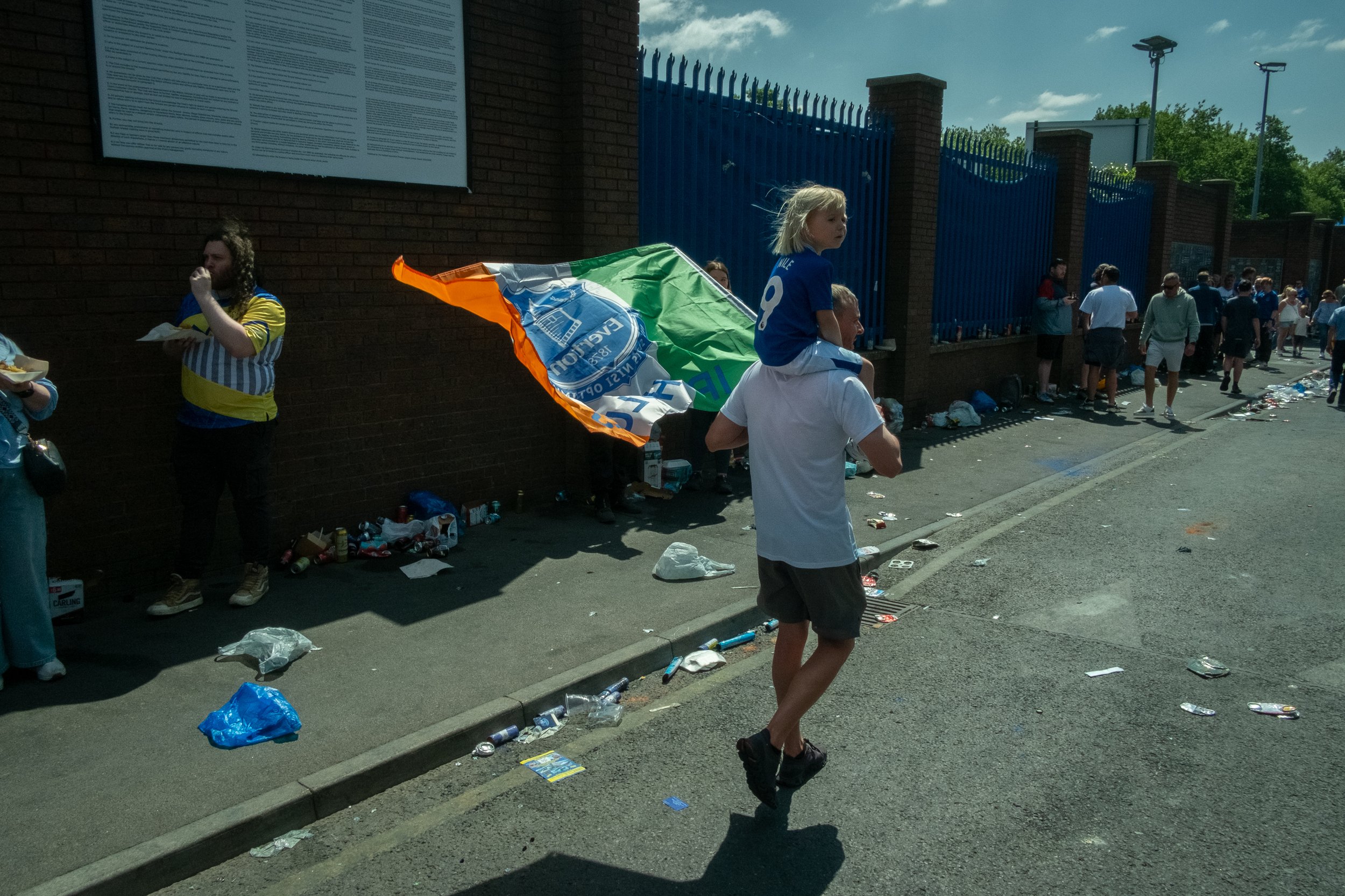 People behind a fence after a sporting event, with debris on the ground and a man carrying a young girl on his shoulders, holding a flag.