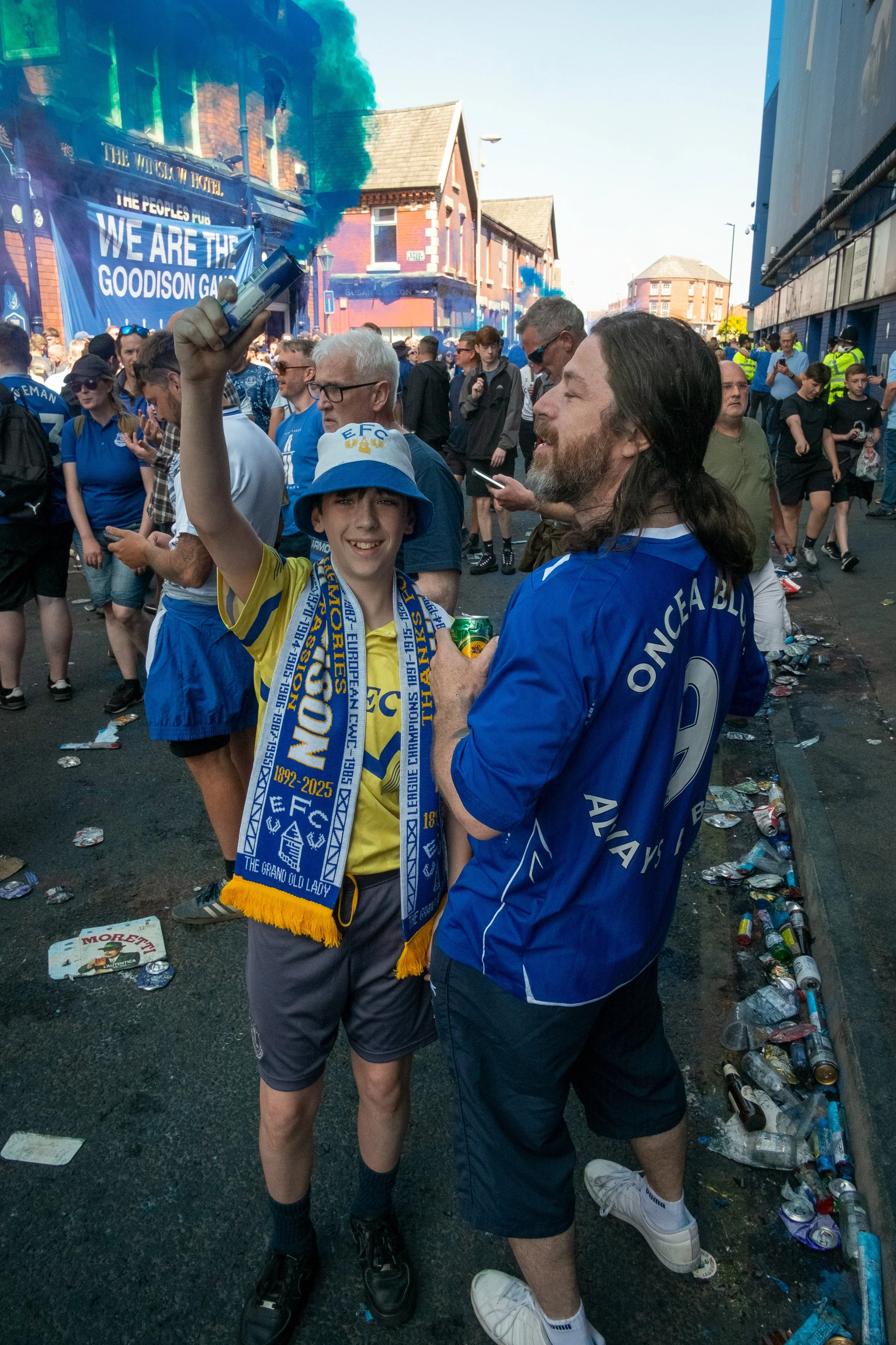 A young boy and an older man, both wearing blue football jerseys and scarves, stand in a crowded street celebrating after a sports event. The boy is holding a smoking flare, and the street is littered with trash.