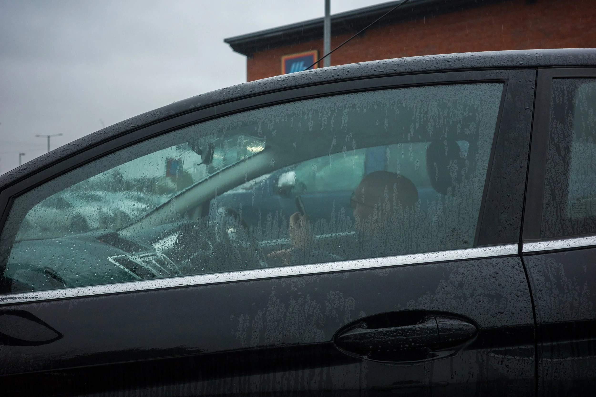 A person sitting inside a black car during rainy weather, with water droplets on the windshield and side window, and a building with a colorful sign in the background.