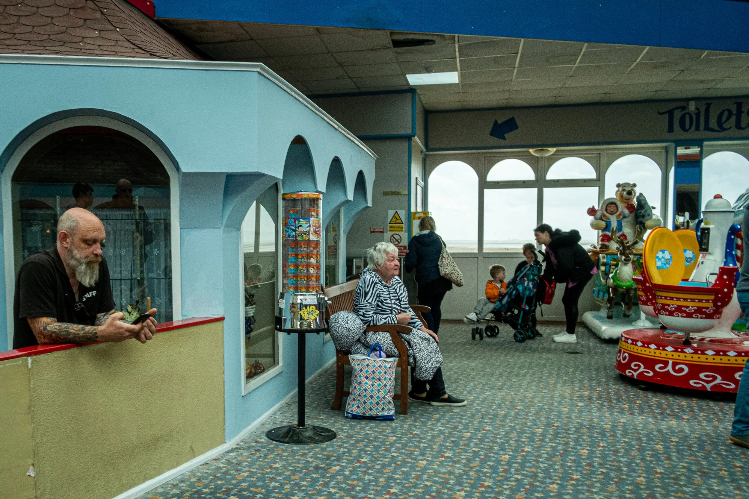 Indoor amusement arcade with people, including a man with tattoos using a phone, an elderly woman sitting on a bench, children, and colorful arcade rides and games.