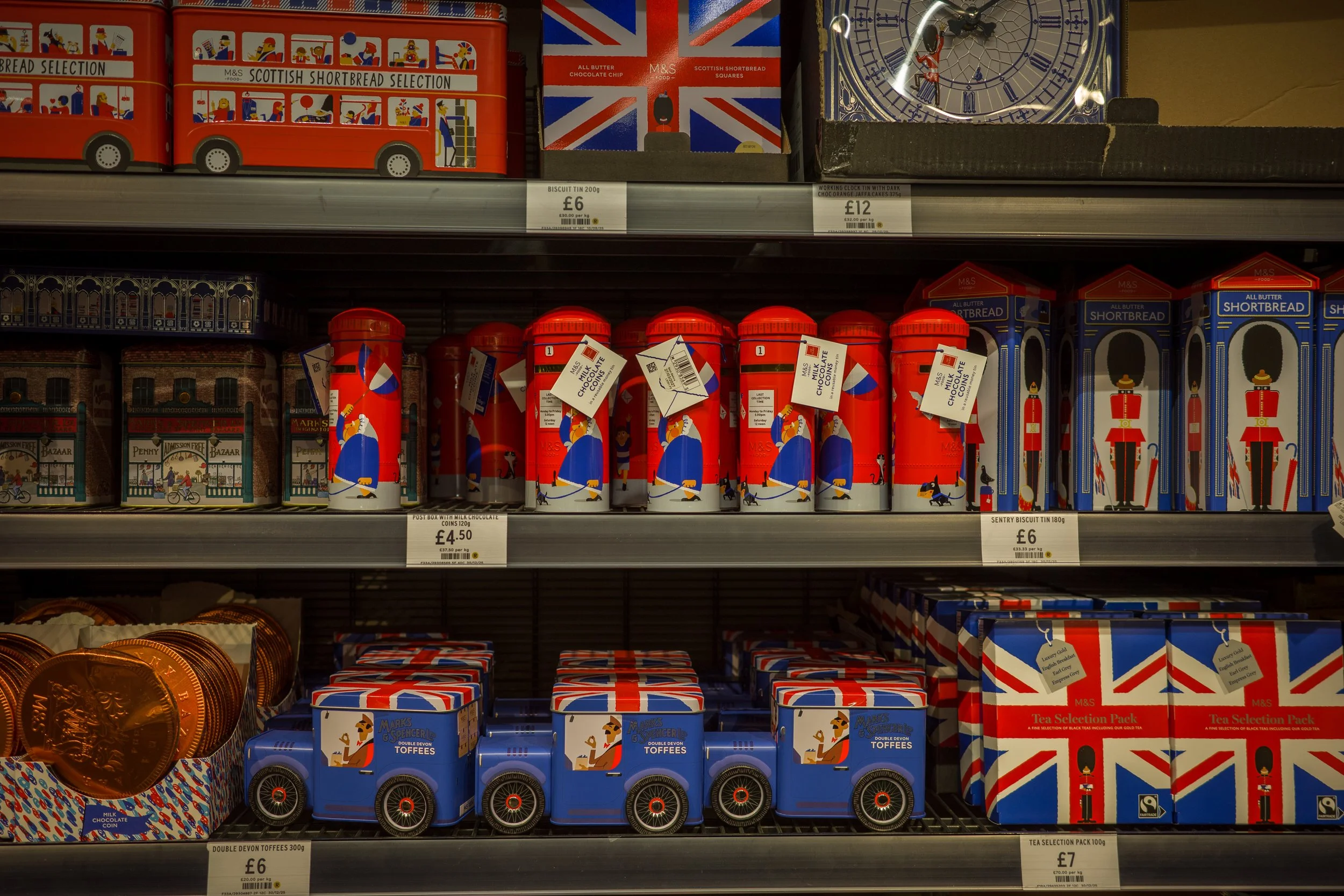 Shelf displaying various British-themed confectionery and tea tins, including Union Jack patterns, British guards, and red double-decker bus designs.