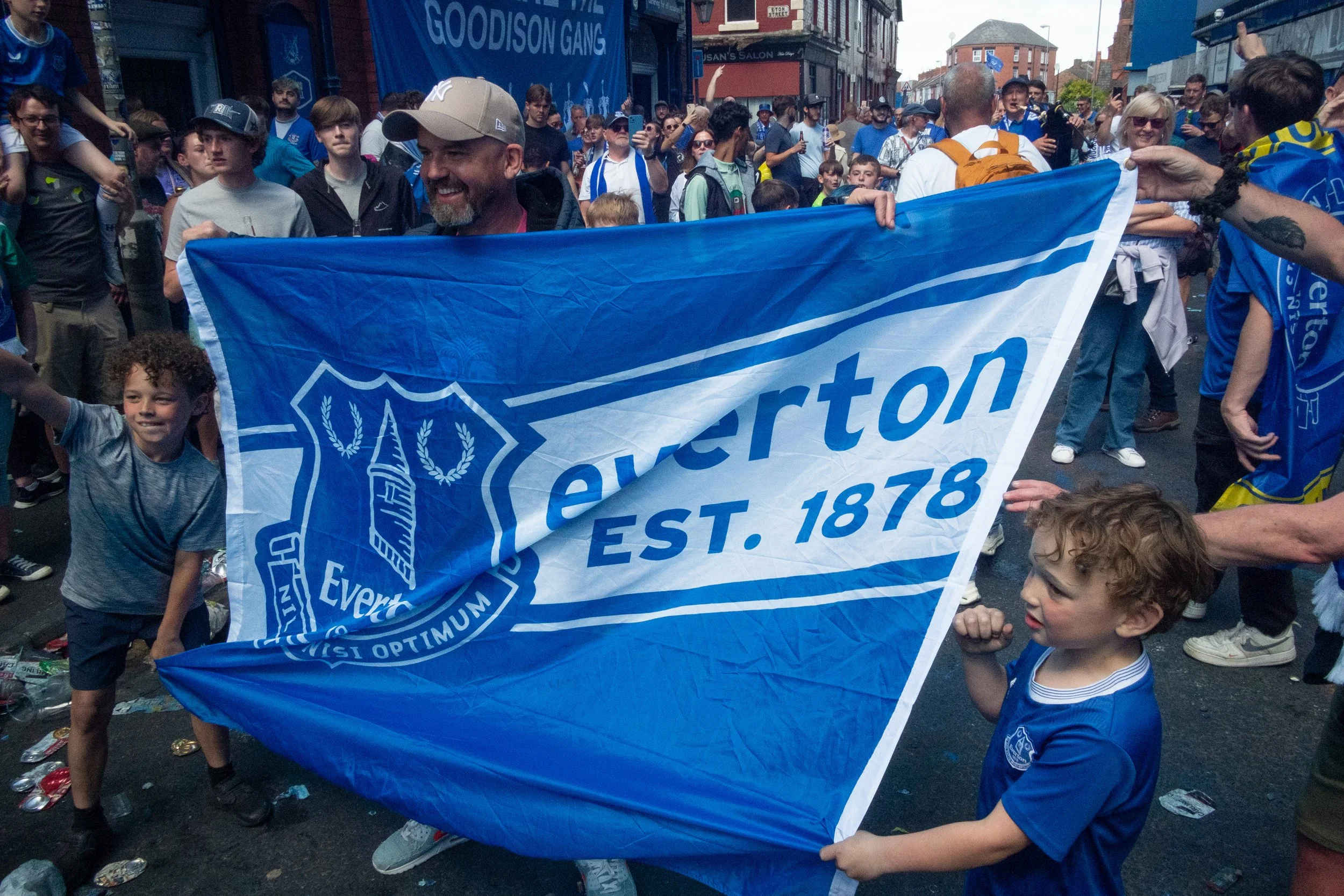 A group of people, including children and adults, holding a large blue Everton Football Club banner that reads 'Everton EST. 1878,' during a parade or celebration.