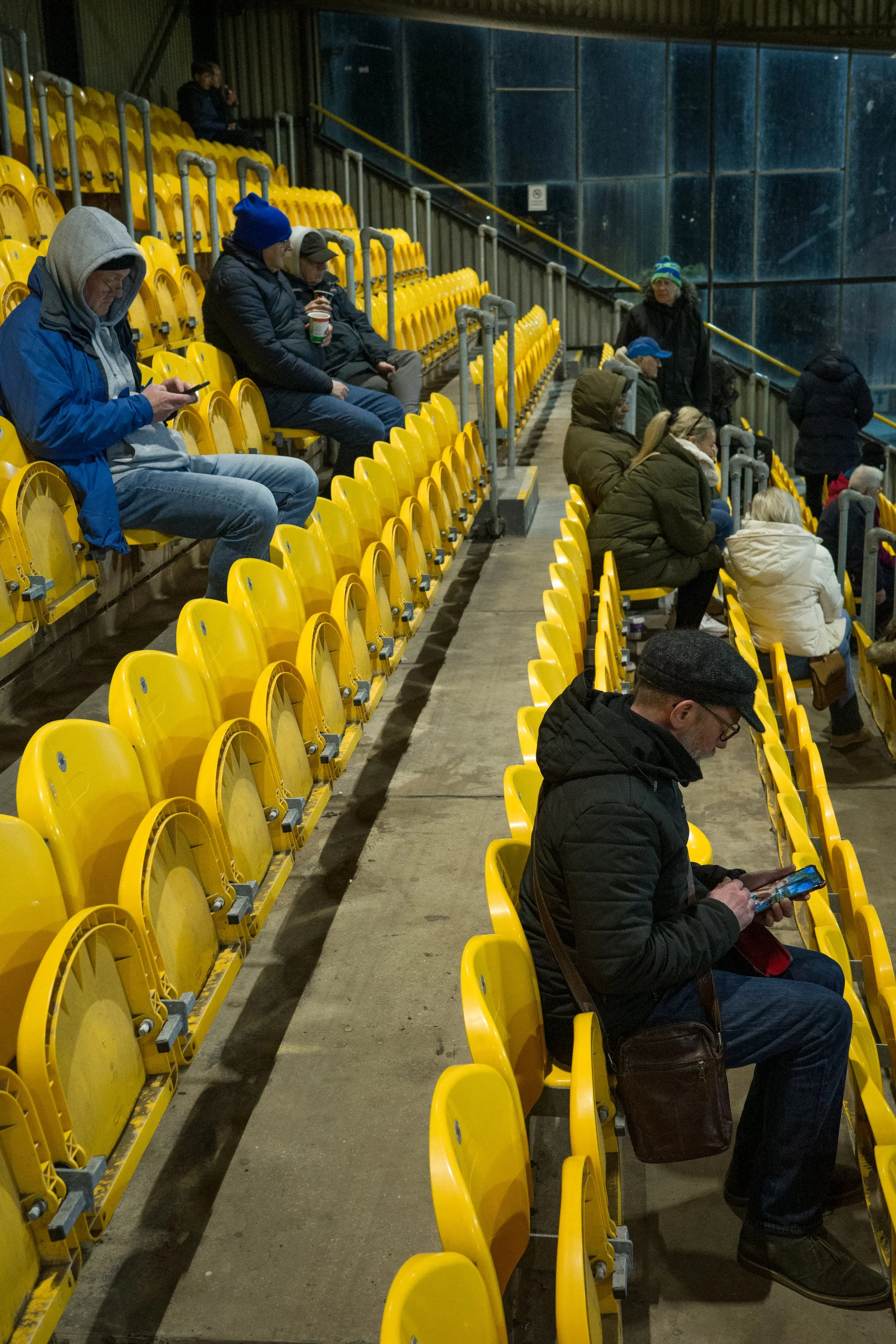People sitting in yellow stadium seats, some using their phones, others holding drinks, inside an indoor sports arena with a glass wall background.