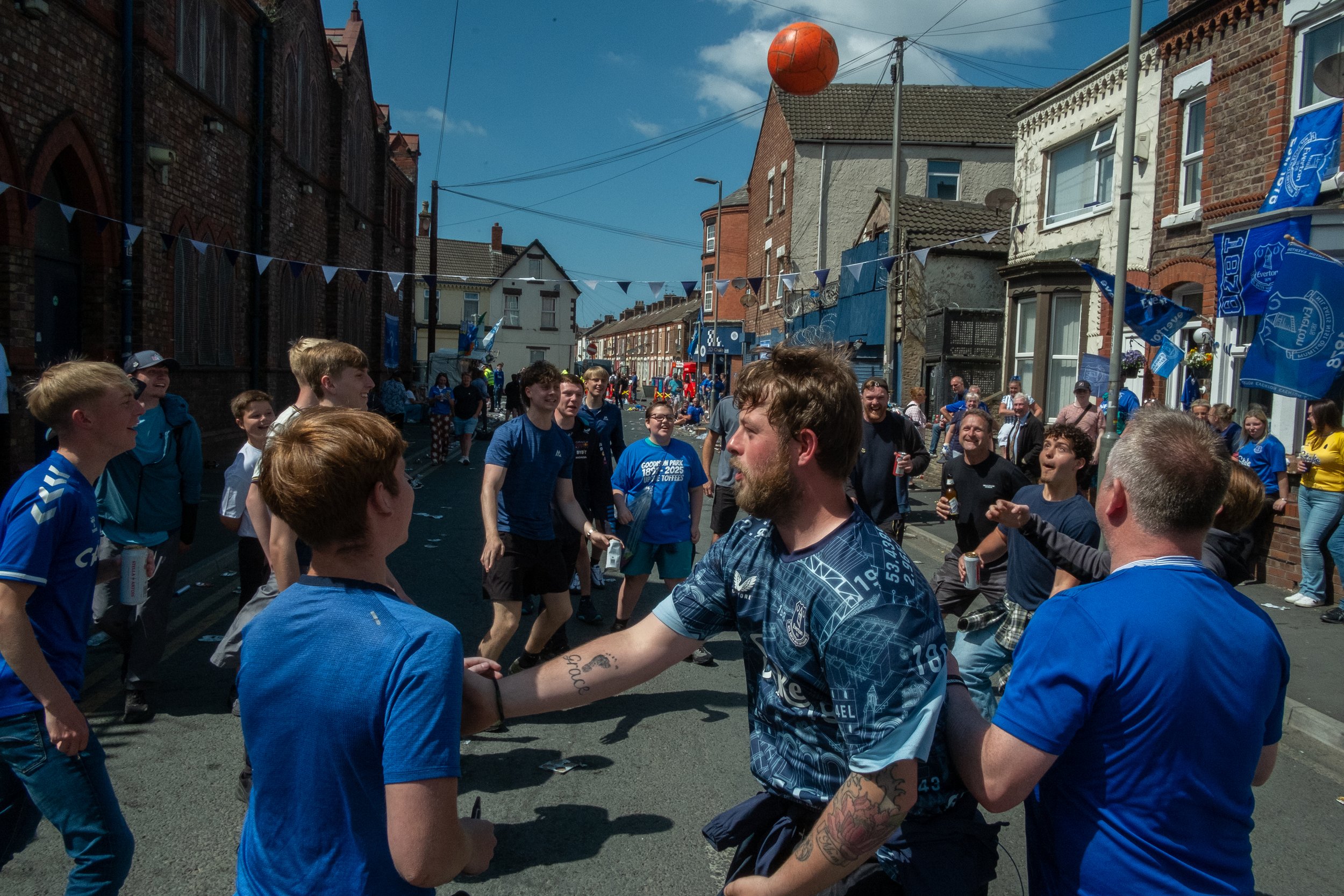 People gathering on a street decorated with blue banners and flags, celebrating with music and dancing under a clear sky, with an orange ball hanging above.