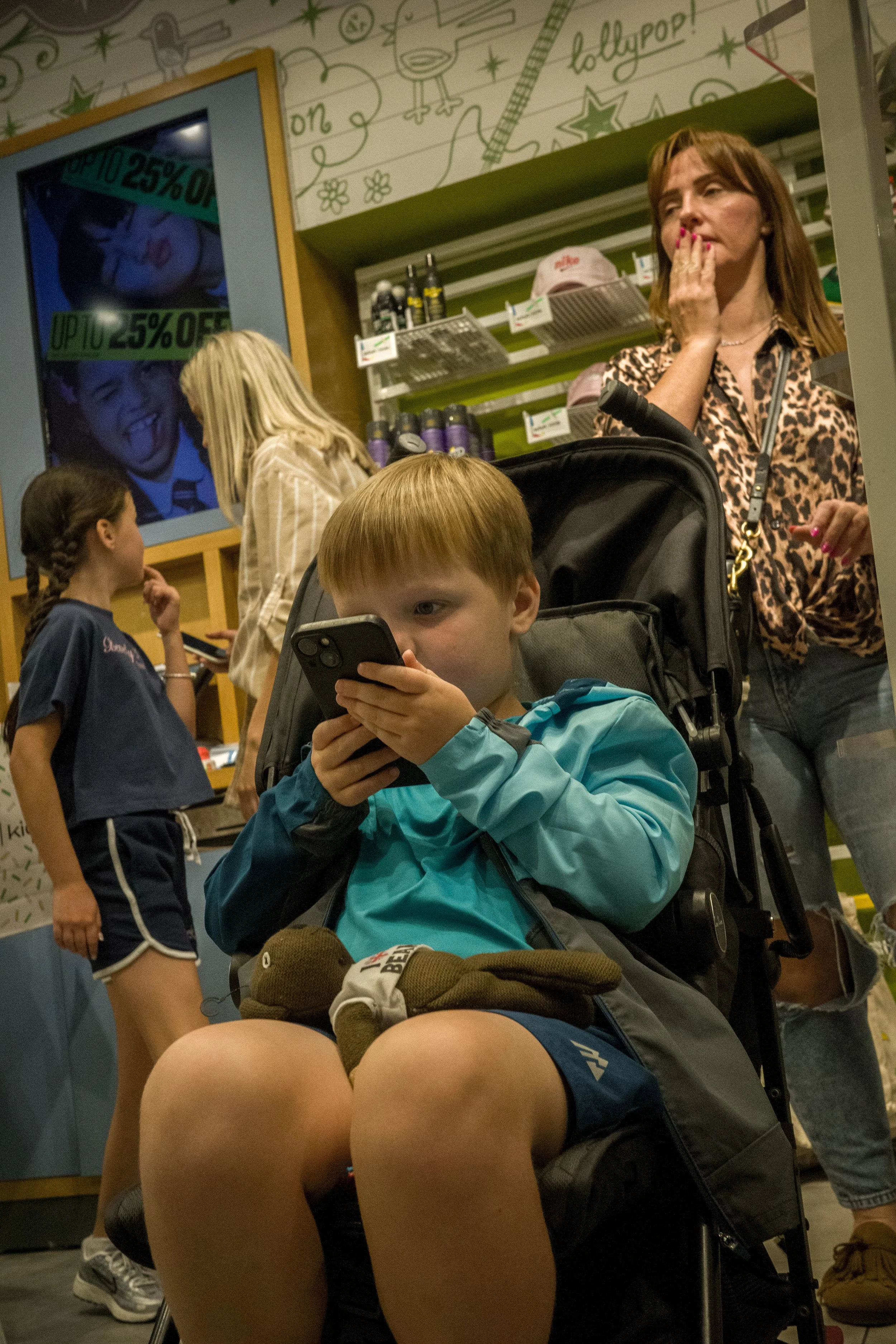 A young boy with red hair sitting in a stroller, wearing a blue jacket, closely looking at a smartphone. Behind him, a woman with brown hair and a leopard print shirt appears to be covering her mouth or yawning. Two other women and a girl are in the 