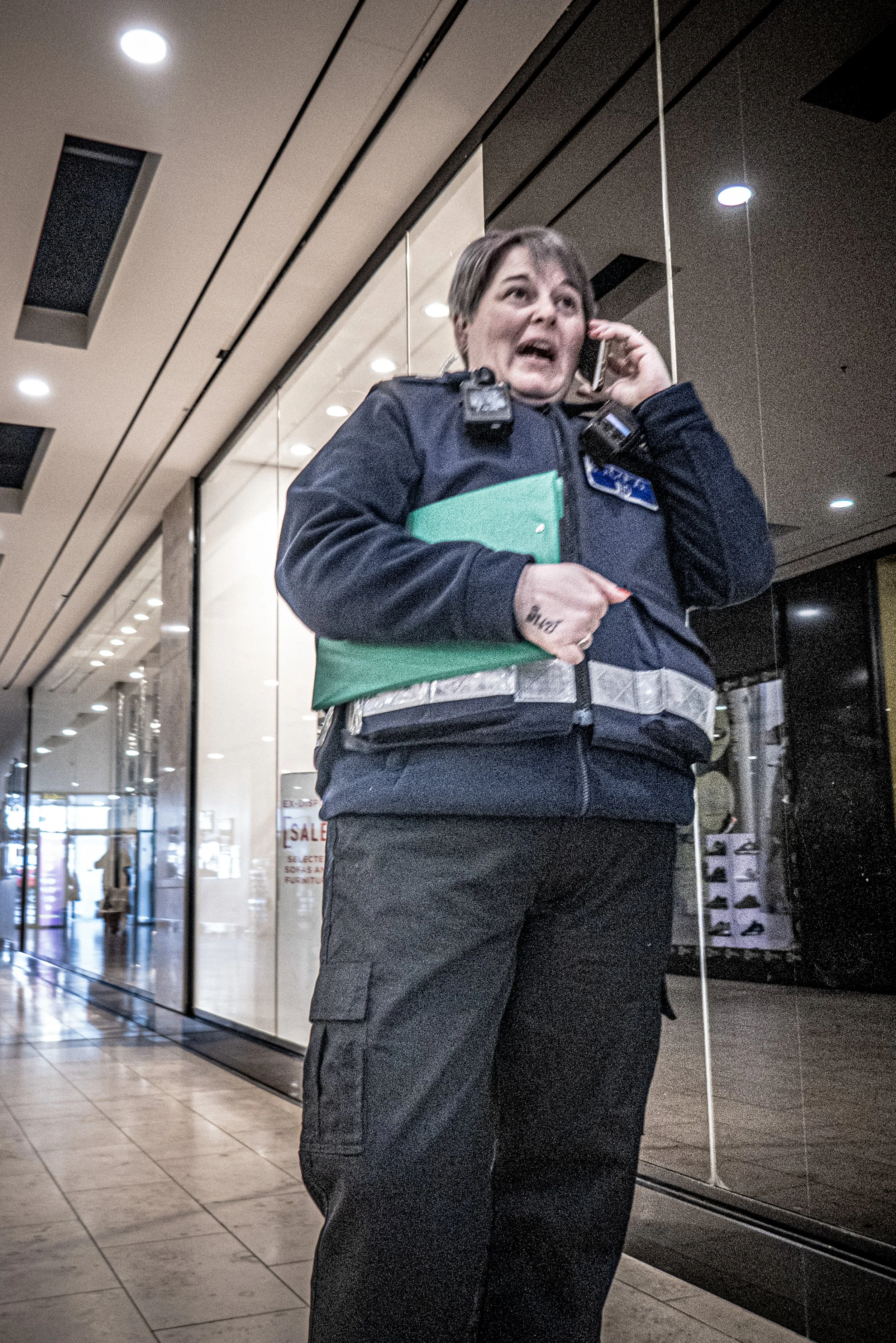 A female security officer or police officer is standing inside a shopping mall or airport, talking on a cell phone, and holding green folders. She is wearing a uniform with a badge and a body camera.